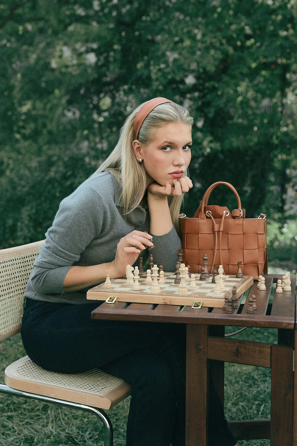Junge Frau spielt Schach im Grünen, trägt grauen Pullover, trägt Kopfband, sitzt an Holztisch, neben Tasche.