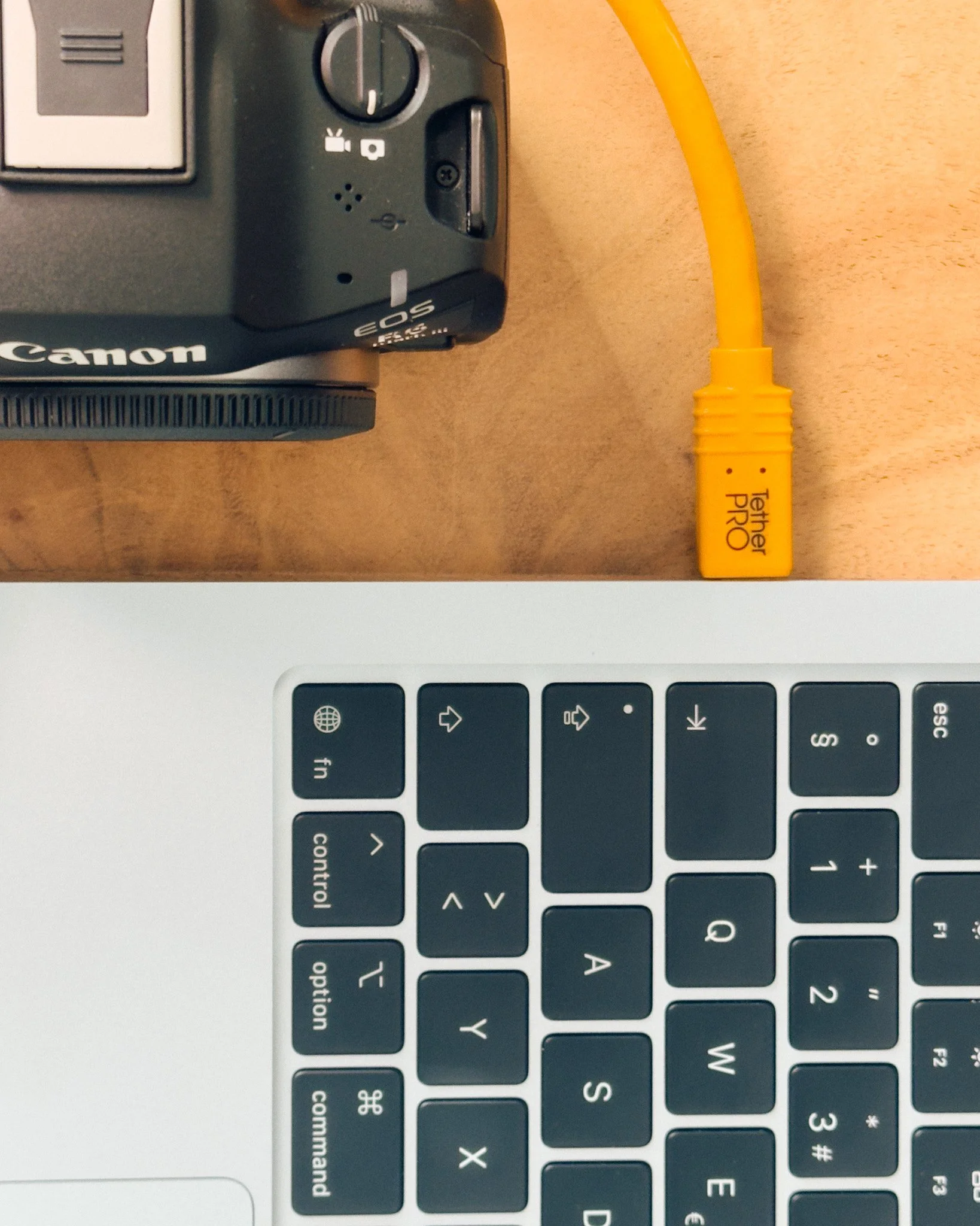 Oberteil einer Kamera und einen gelben Netzwerk-Adapter auf einem Holztisch, darunter Teil eines Laptops mit Tastatur.