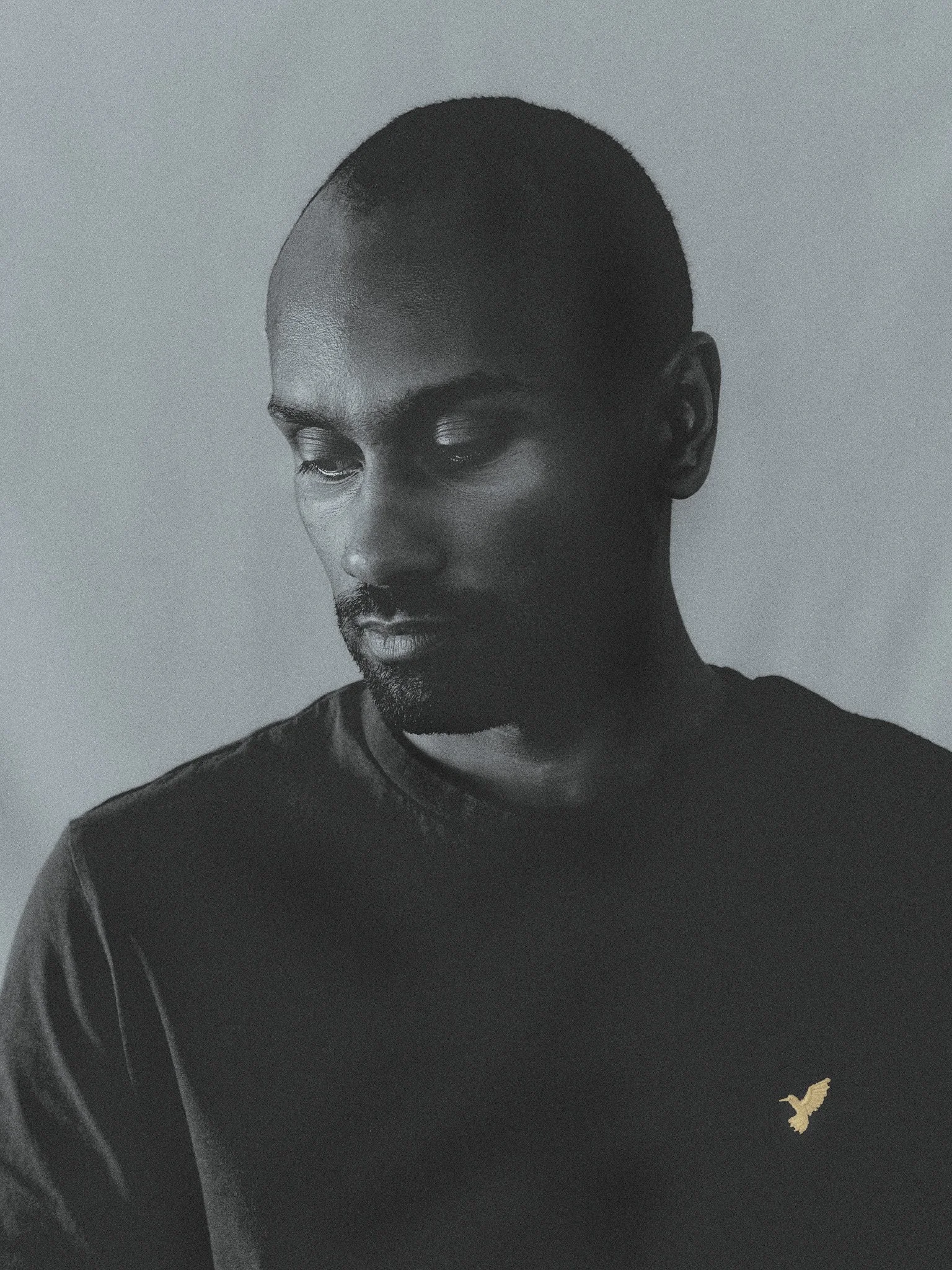 Schwarz-weiß-Foto eines Mannes mit kurz geschnittenem Haar und dunkler Haut, der nach unten blickt, trägt ein schwarzes T-Shirt mit einem kleinen goldenen Vogel-Logo auf der Brust.