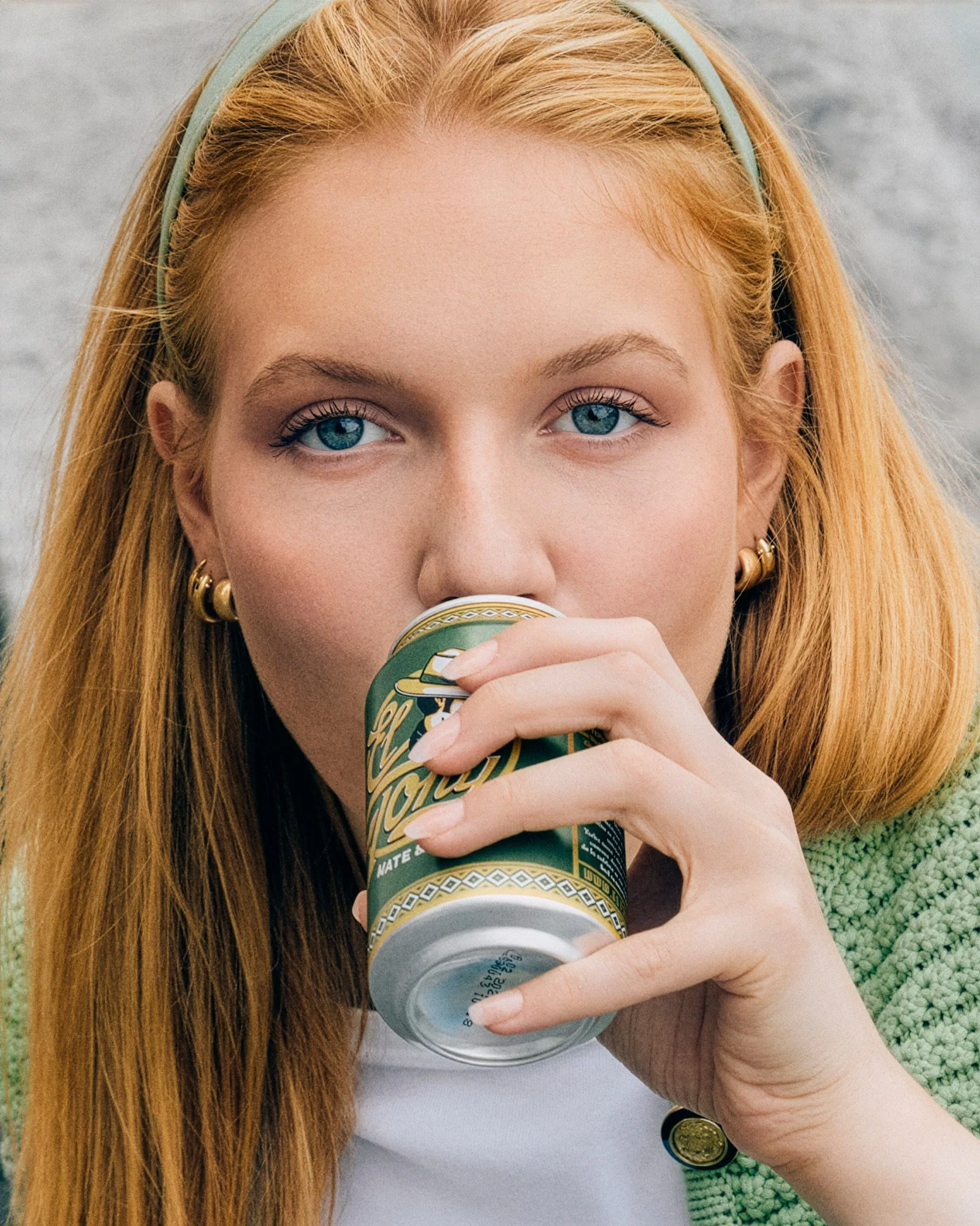 Nahaufnahme eines Mädchens mit roten Haaren und blauen Augen, das eine Dose trinkt. Das Mädchen trägt goldene Ohrringe und einen grünen Pullover.