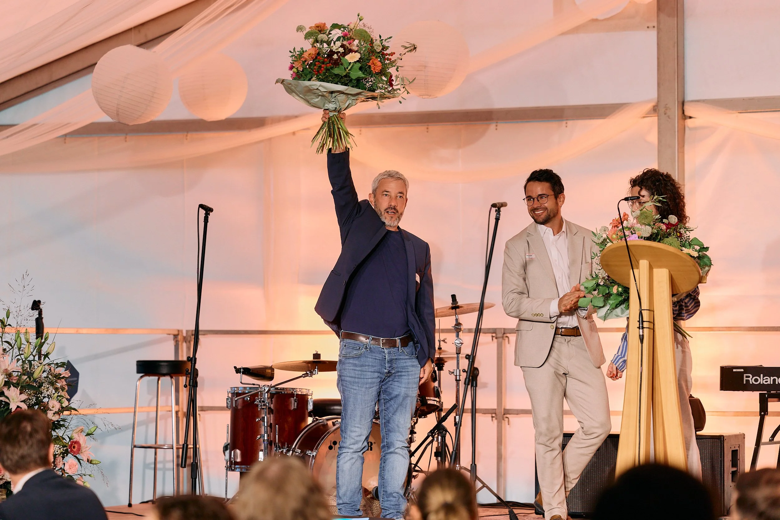 Ein Mann mit grauem Bart hält einen Blumenstrauß hoch, neben ihm stehen ein Mann in Brille und Krawatte sowie eine Frau mit Locken und Brille, die Blumen hält. 100 Jahre Gymnasiums Friedberg Gossau SG.
