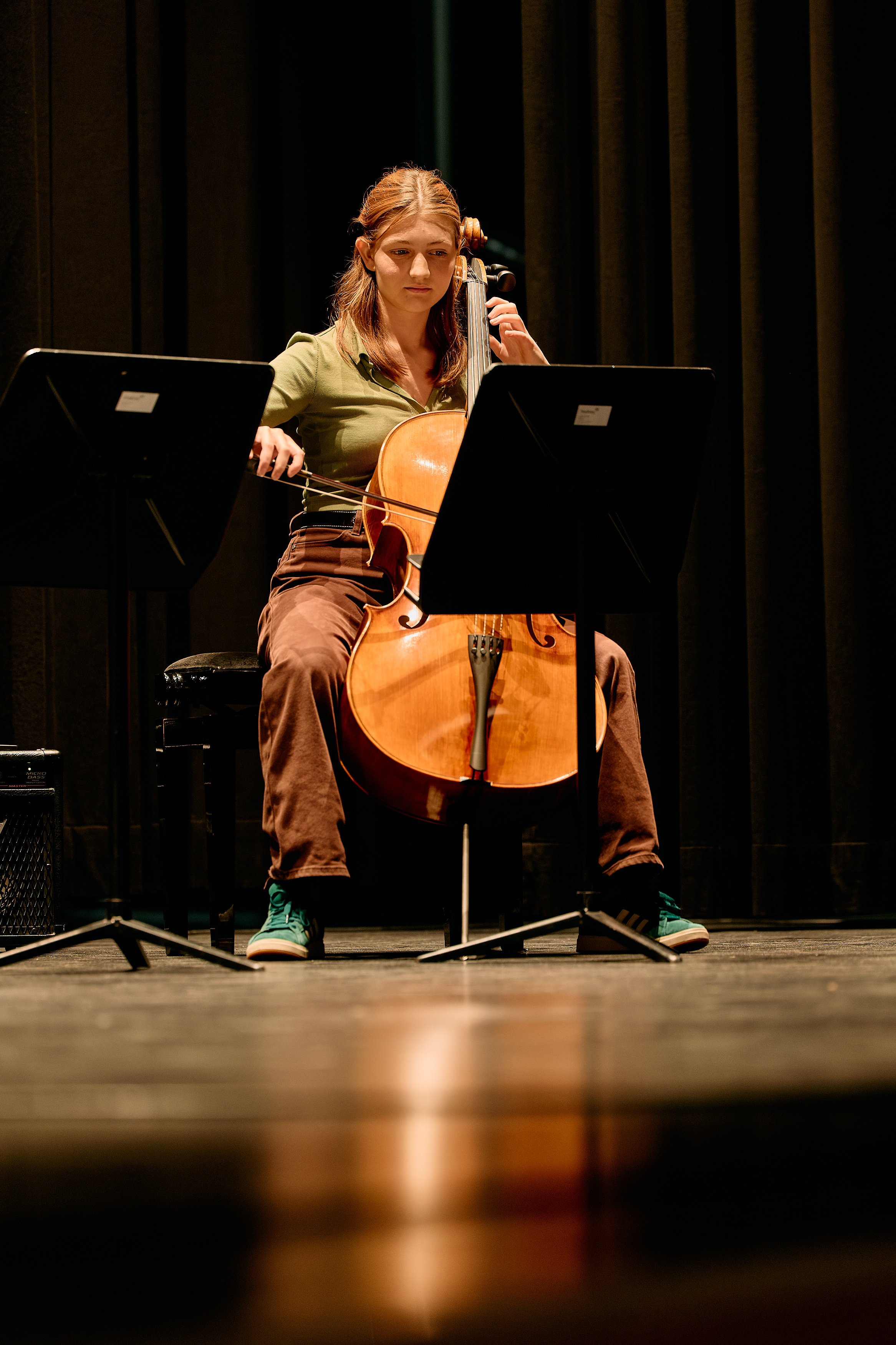 Eine junge Frau spielt auf der Bühne Cello, umgeben von Musikständern, in einem dunklen Raum.