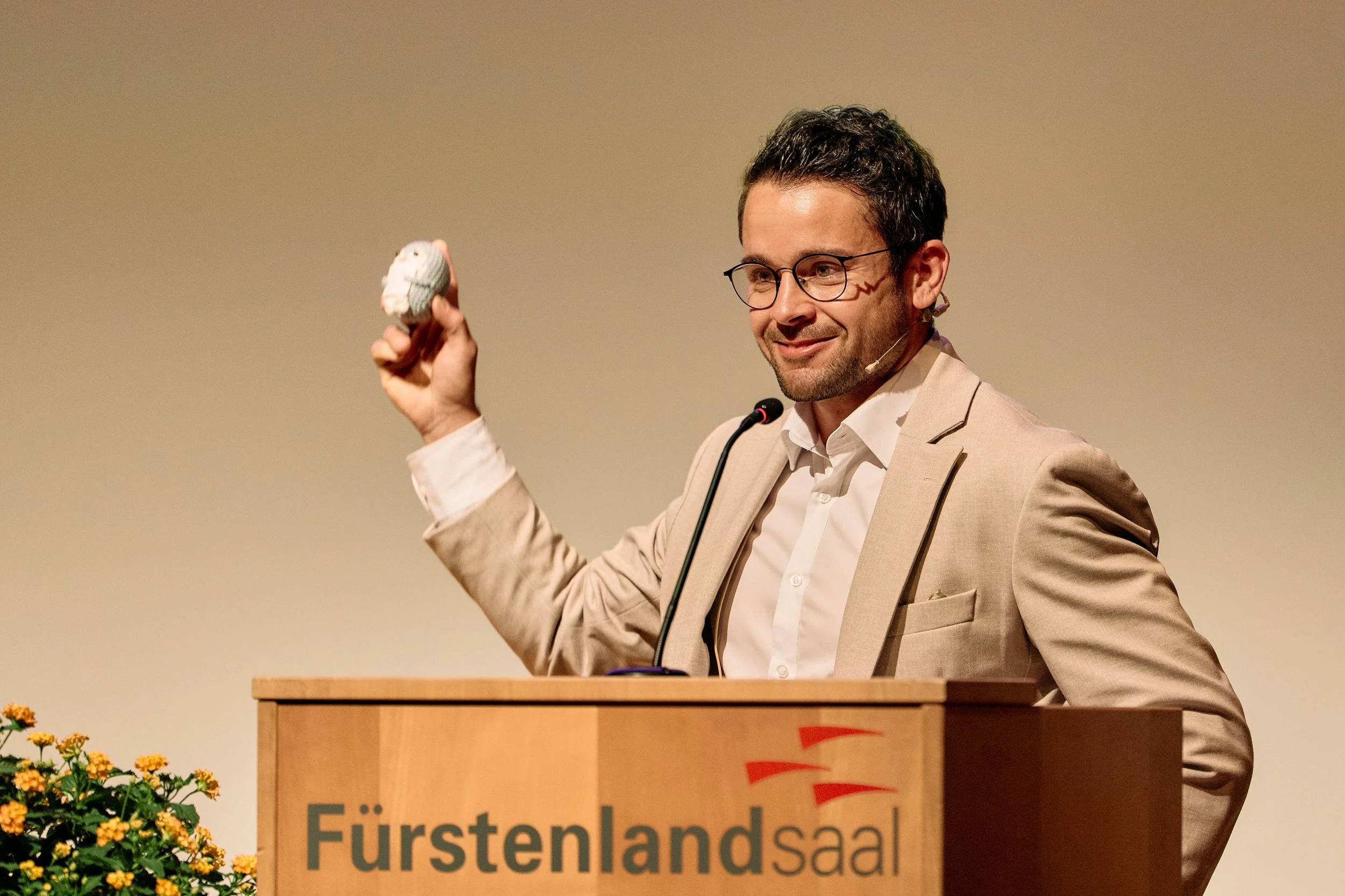 Mann steht an einem Rednerpult mit einem Mikrofon und hält eine kleine Figur in der Hand. Er trägt eine beige Jacke, weiße Hemd und hat eine Brille. Im Hintergrund ist eine neutrale Wand, und es sind gelbe Blumen im Vordergrund sichtbar.