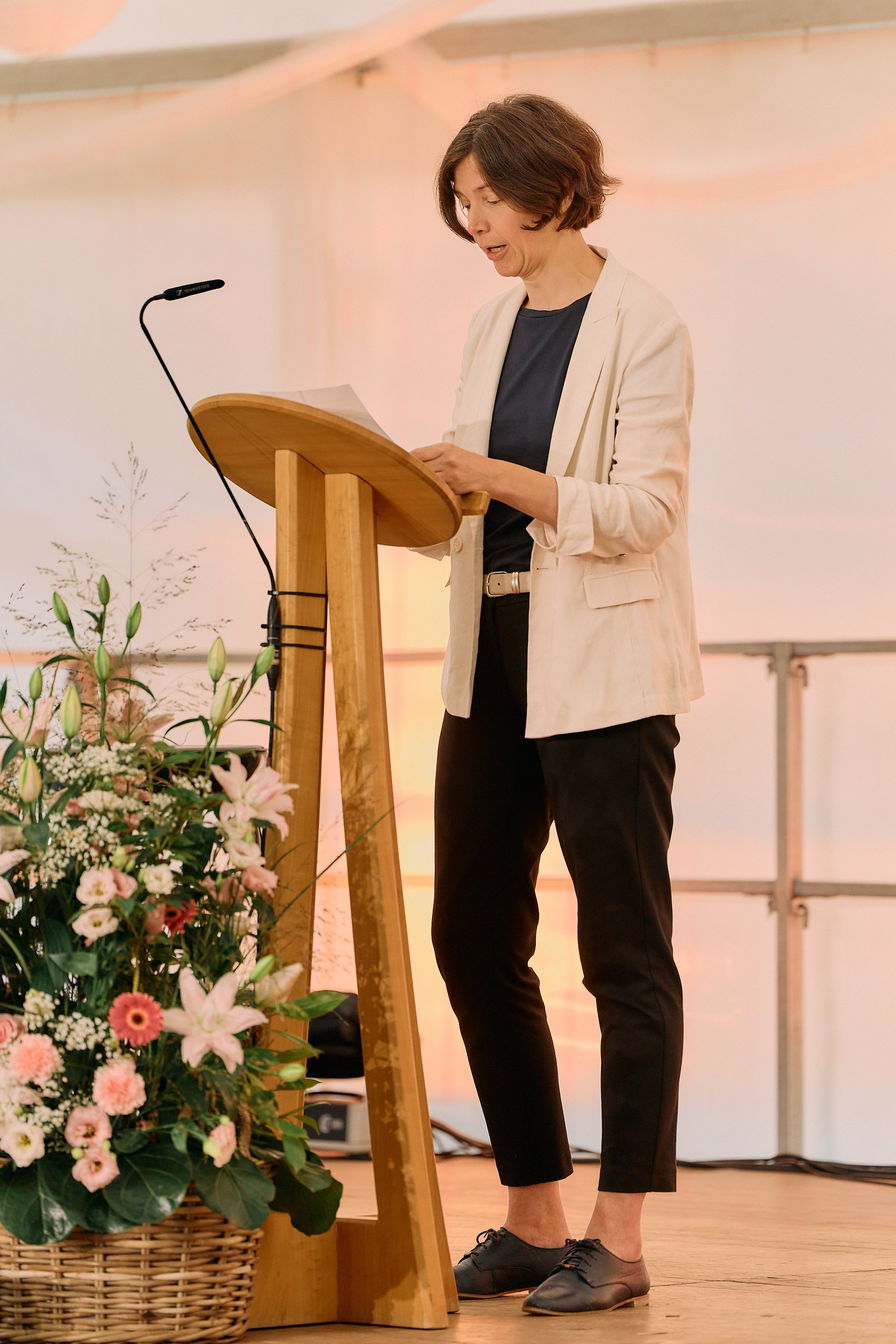 Fotograf & Videograf Wil SG | Businessfotografie Ostschweiz Daniel Dorrer Frau steht an einem Holzpodest und liest aus Papier, umgeben von Blumen, in einem Raum mit heller Wand. 100 Jahre Gymnasiums Friedberg Gossau SG. Bettina Surber