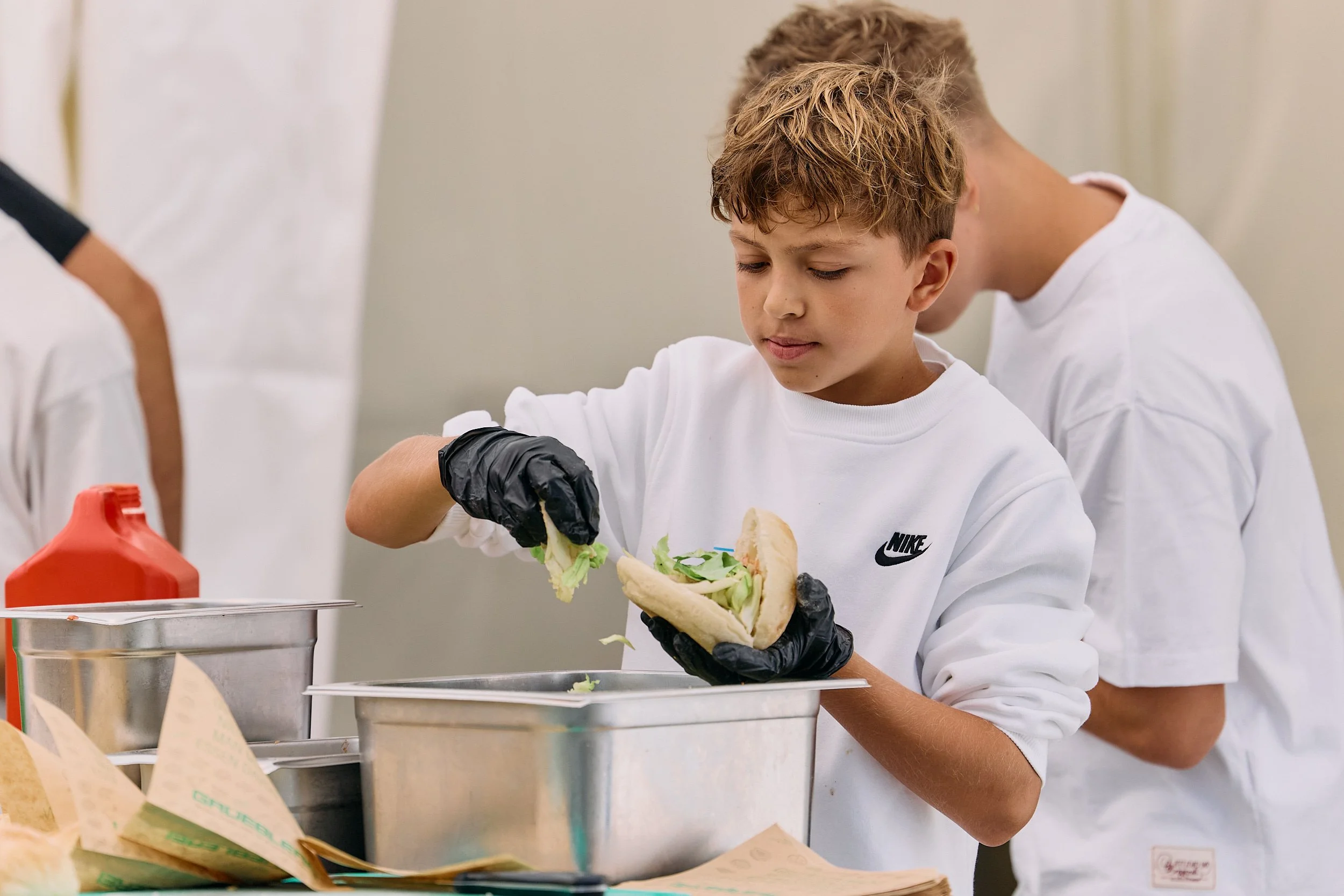 Ein Junge trägt schwarze Handschuhe und hält ein Sandwich mit Salat, während sich im Hintergrund weitere Menschen in weißen Kochjacken befinden. 100 Jahre Gymnasiums Friedberg Gossau SG.