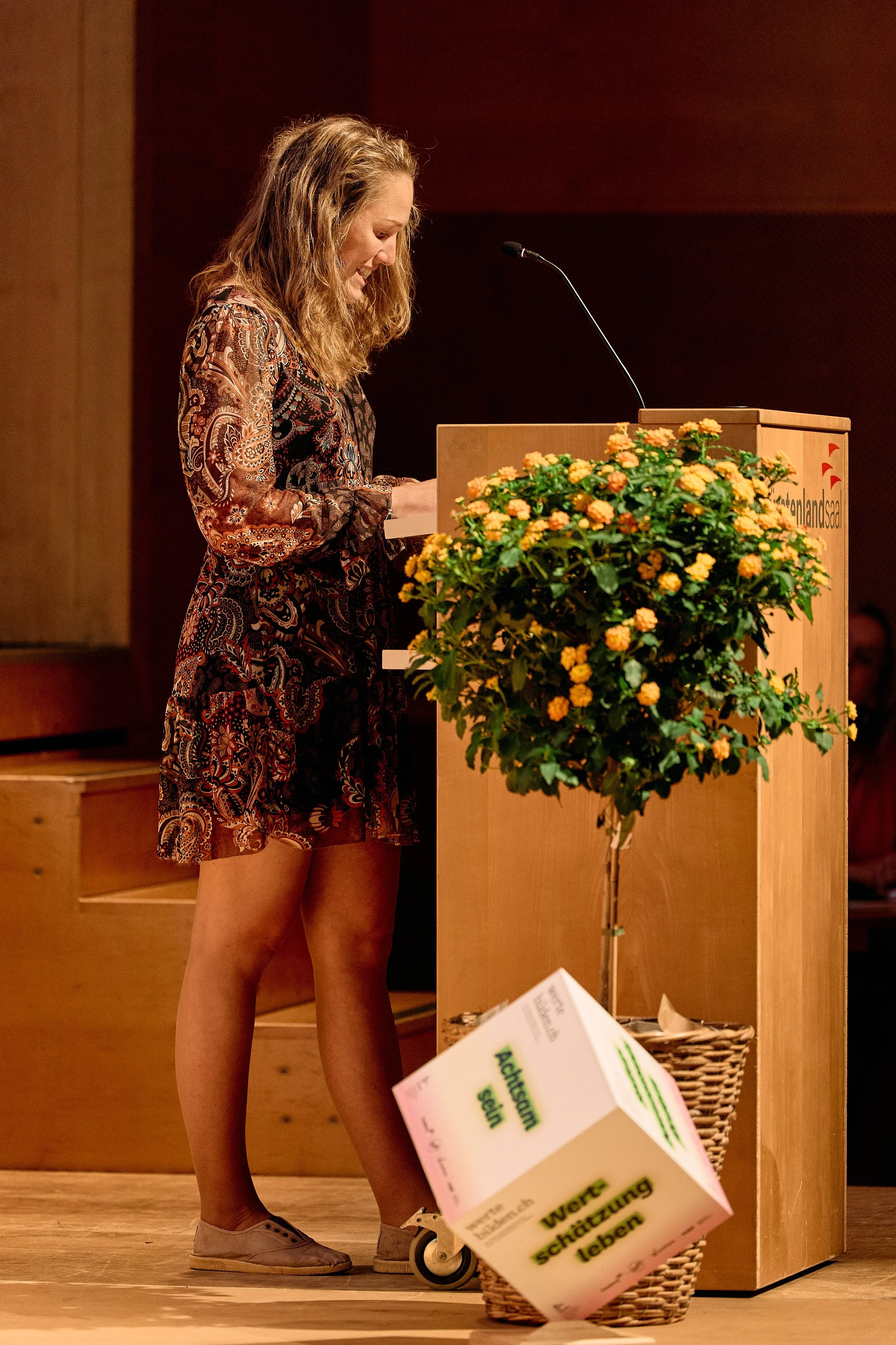 Eine Frau steht bei einer Veranstaltung hinter einem Rednerpult, umgeben von Blumen, und liest oder spricht in ein Mikrofon.