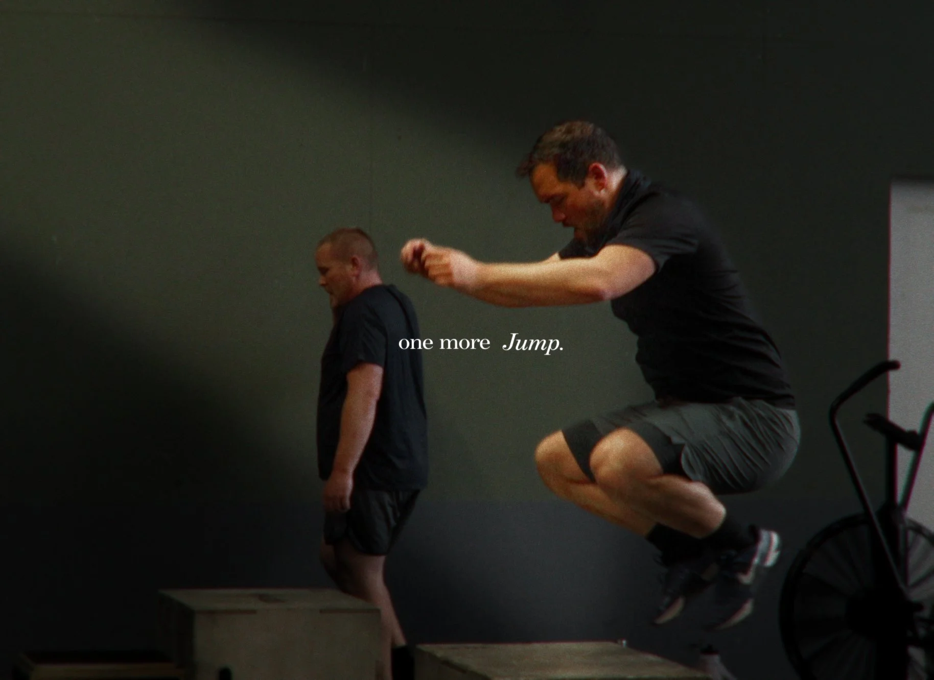 Zwei Männer trainieren im Fitnessstudio. Einer springt auf eine Box, der andere steht auf dem Boden. Der Text auf dem Bild lautet 'one more Jump.'