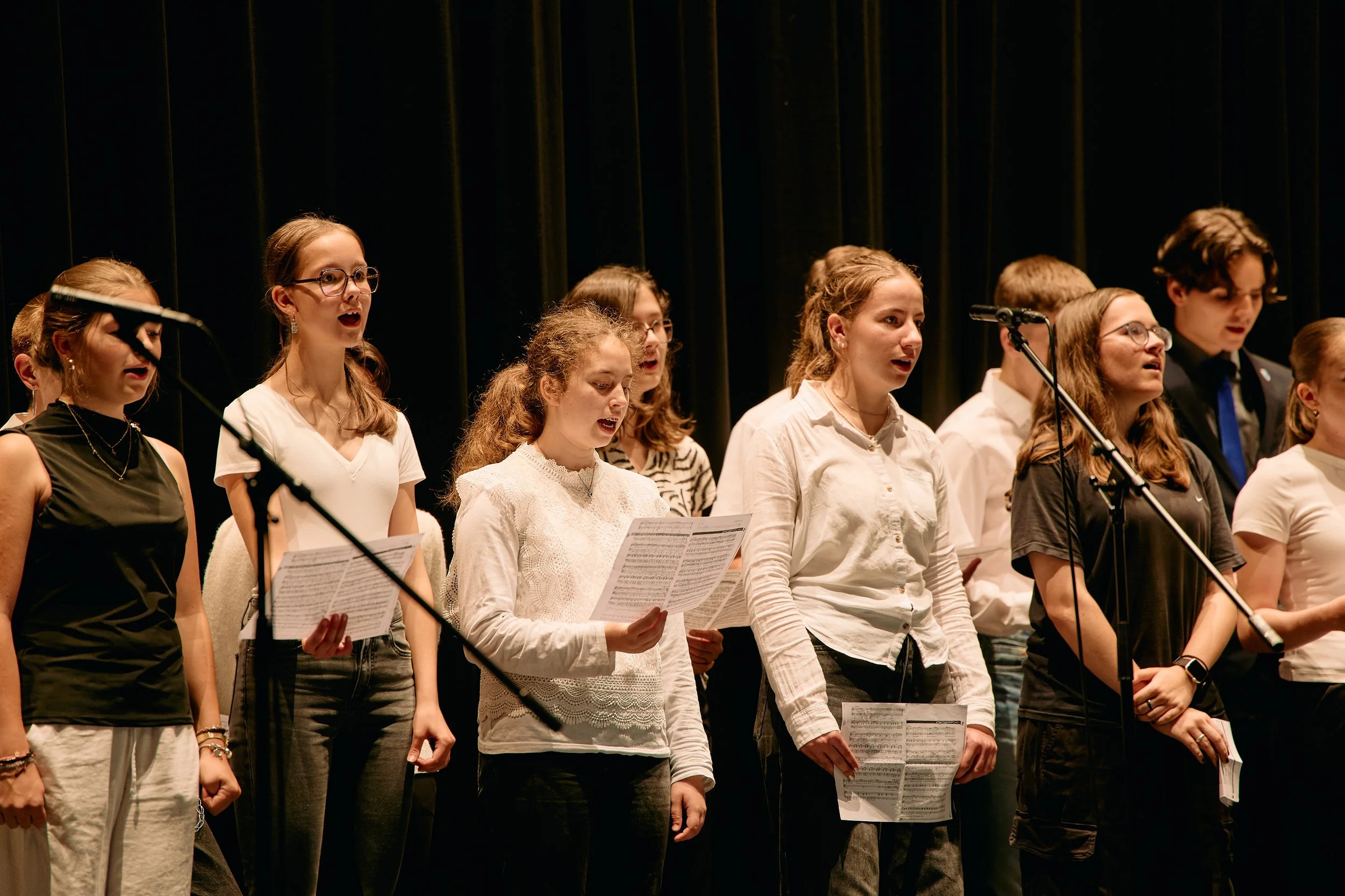 Jugendchor singt auf einer Bühne mit Notenblättern in der Hand, im Hintergrund Dunkler Vorhang.