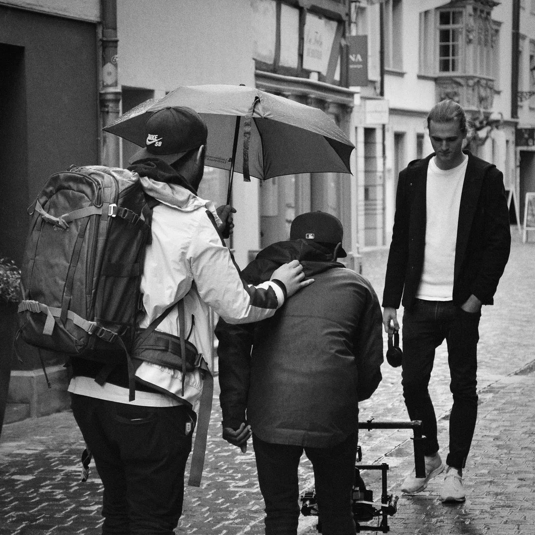Drei Männer auf einer Straße, einer hält einen schwarzen Regenschirm, ein Filmemacher mit Kamera, andere Person schaut auf den Boden.