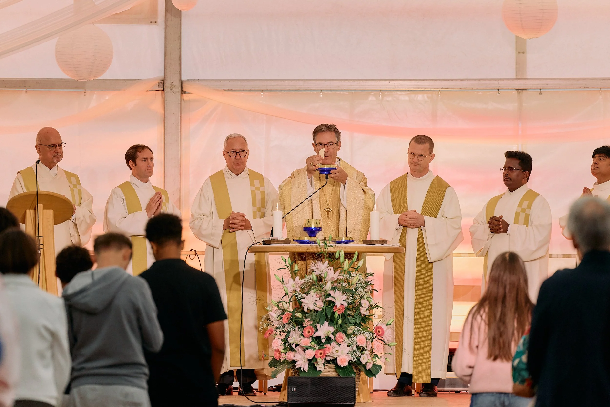 Priester bei einer katholischen Messe, die vor einem Altar mit Blumen steht, in einer Kirche oder Festhalle, mit Gläubigen im Gebet. 100 Jahre Gymnasiums Friedberg Gossau SG. Bischof Beat Grögli