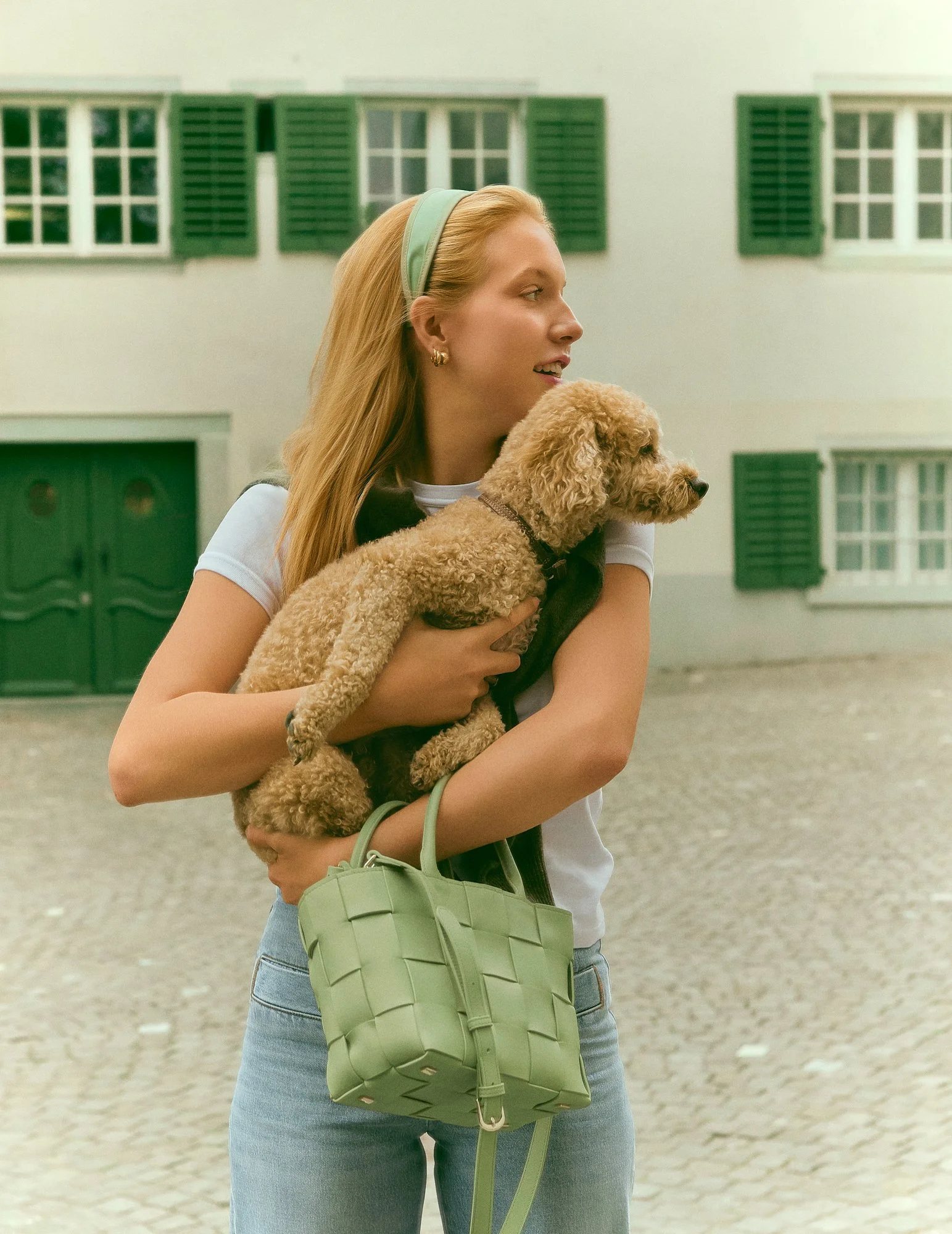Eine junge Frau mit blonden Haaren trägt einen grünen Einkaufskorb und hält einen kleinen braunen Hund. Sie steht vor einer weißen Wand mit grünen Fensterläden auf der Straße.