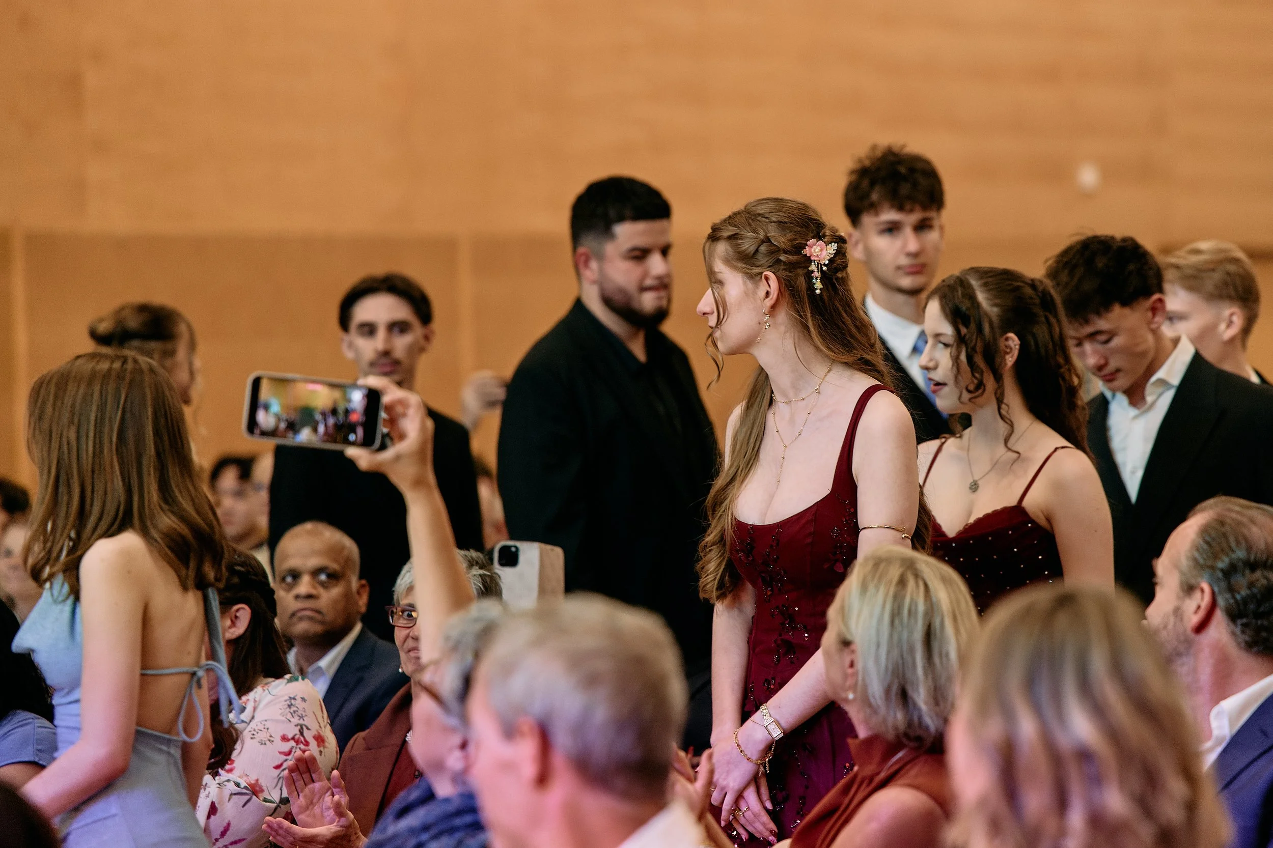 Junge Menschen in formeller Kleidung bei einer Veranstaltung, eine Frau in einem roten Kleid im Mittelpunkt, andere Personen im Hintergrund, einige werden gefilmt oder fotografiert.
