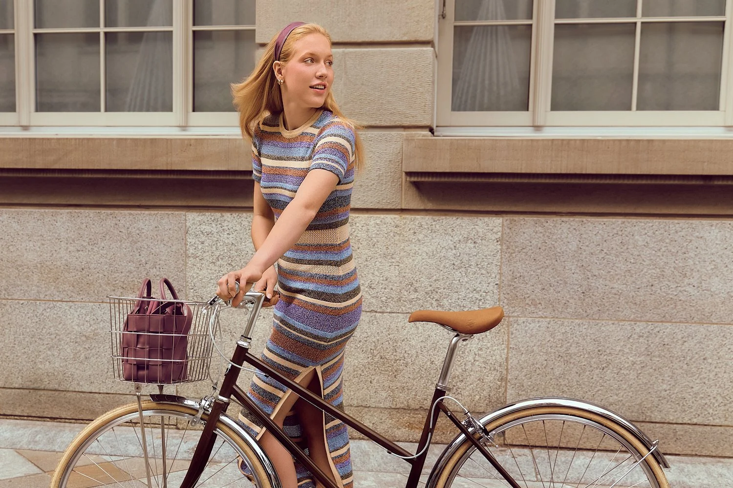 Eine junge Frau mit langem, blondem Haar in einem bunten, gestreiften Kleid steht neben einem Fahrrad mit einem Korb, der eine pinke Tasche trägt, vor einer Steinmauer und großen Fenstern.
