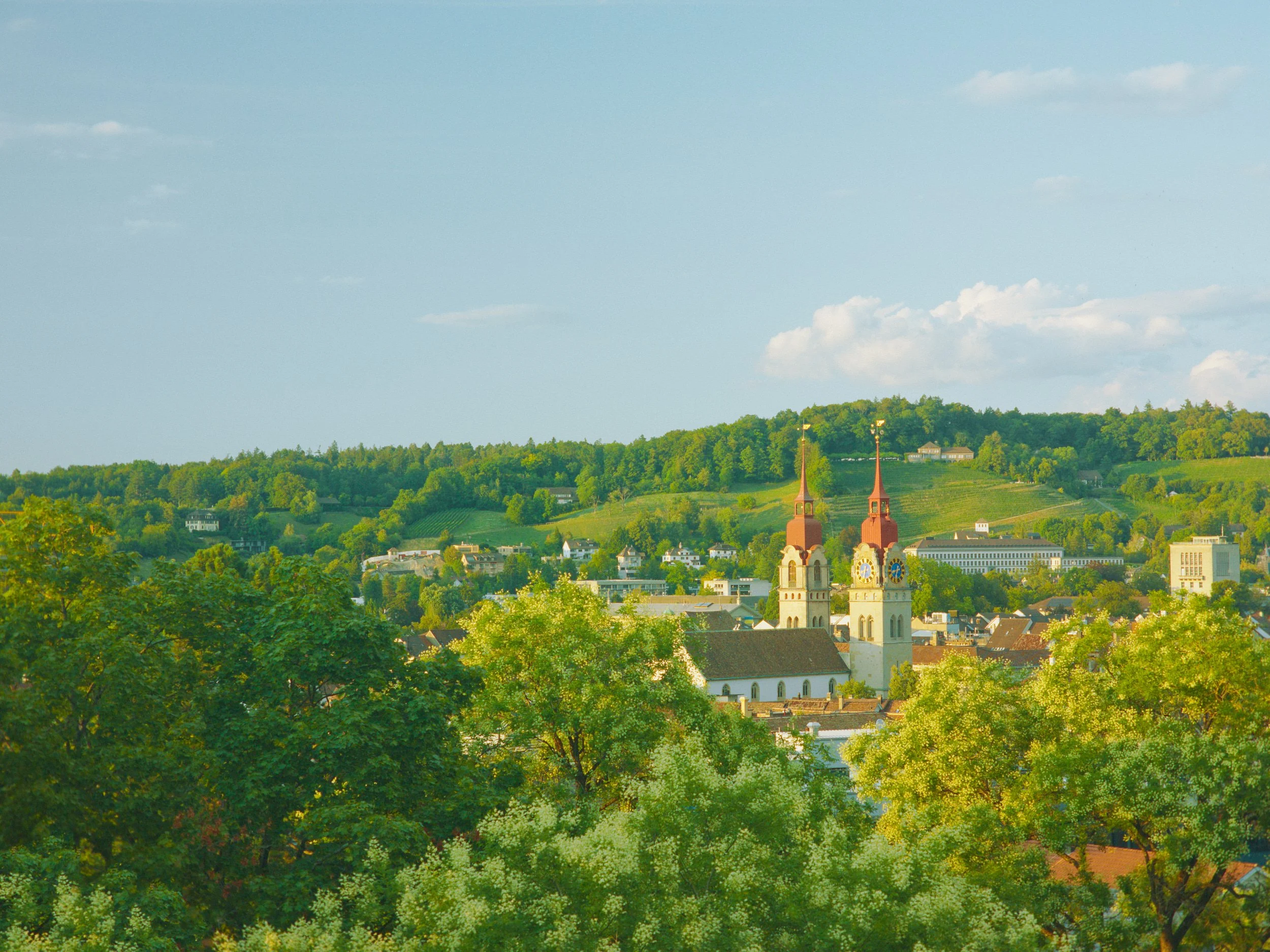 Ein Panorama-Bild einer Stadt inmitten grüner Hügeln mit zwei Kirchtürmen im Vordergrund, blauer Himmel mit einigen Wolken.