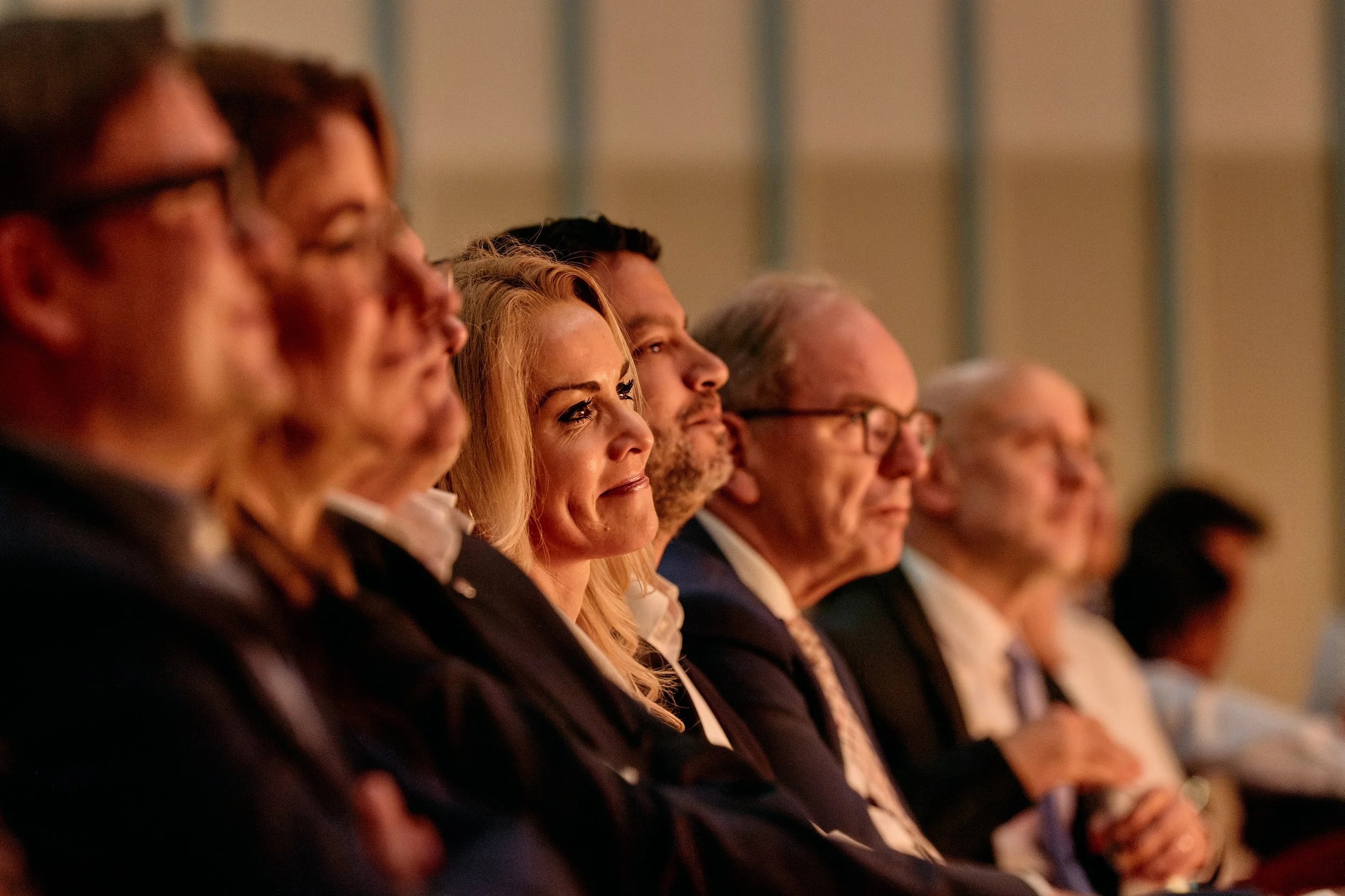Linie von Menschen in formeller Kleidung, die einem Vortrag oder einer Präsentation lauschen, bei einer Veranstaltung im Innenraum.