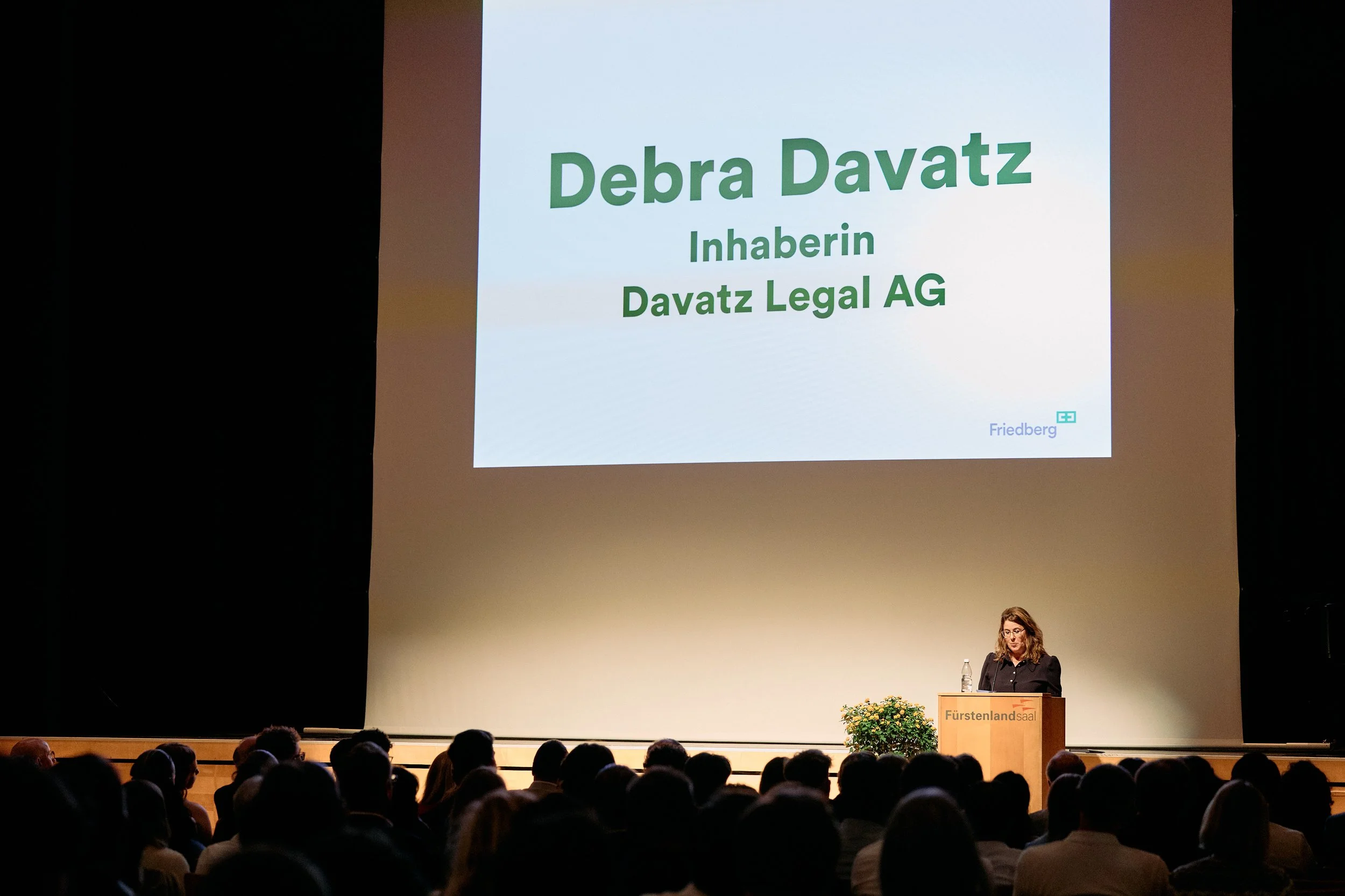 Eine Frau steht an einem Rednerpult vor einem Publikum. Im Hintergrund ist eine große Leinwand mit dem Text 'Debra Davatz, Inhaberin Davatz Legal AG'.