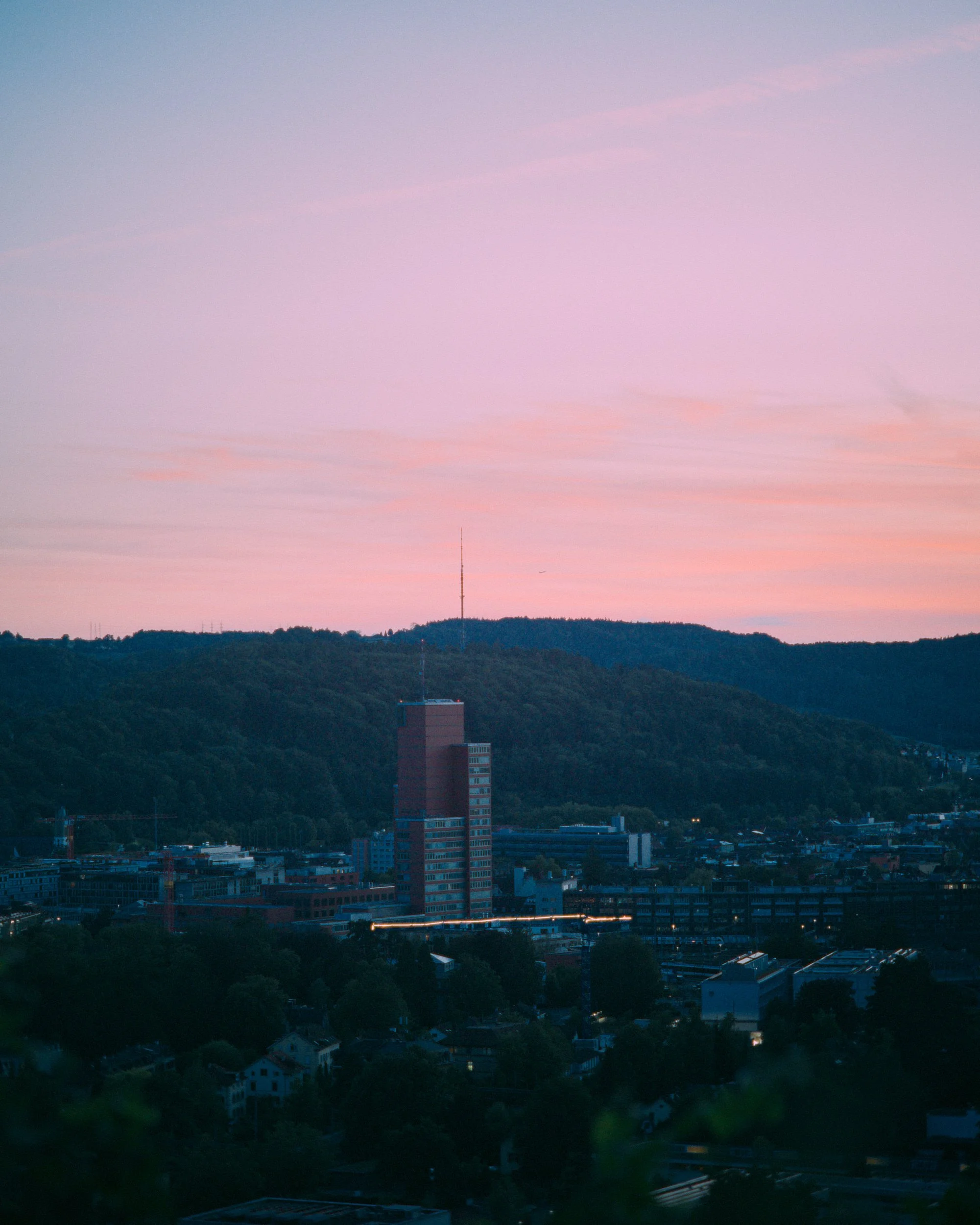 Stadtansicht bei Sonnenuntergang mit Himmel in Pastellfarben, im Vordergrund Gebäude und Bäume, im Hintergrund bewaldete Hügel und eine Antennen- oder Funkturmspitze.