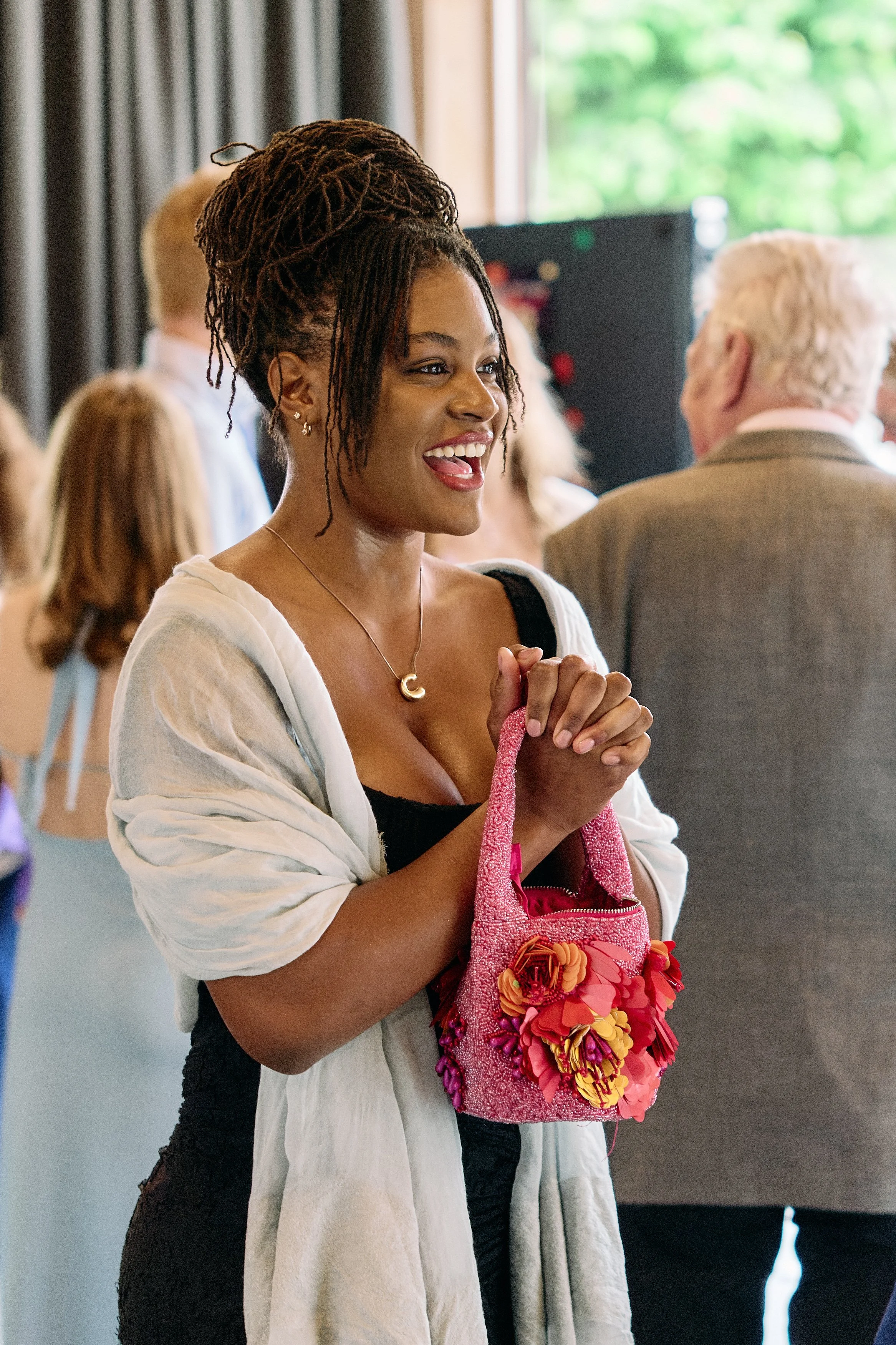 Eine junge schwarze Frau mit dreadlocks, die eine pinke Tasche mit Blumen hält, ist in einem Raum mit anderen Menschen. Sie lächelt und sieht erfreut aus.