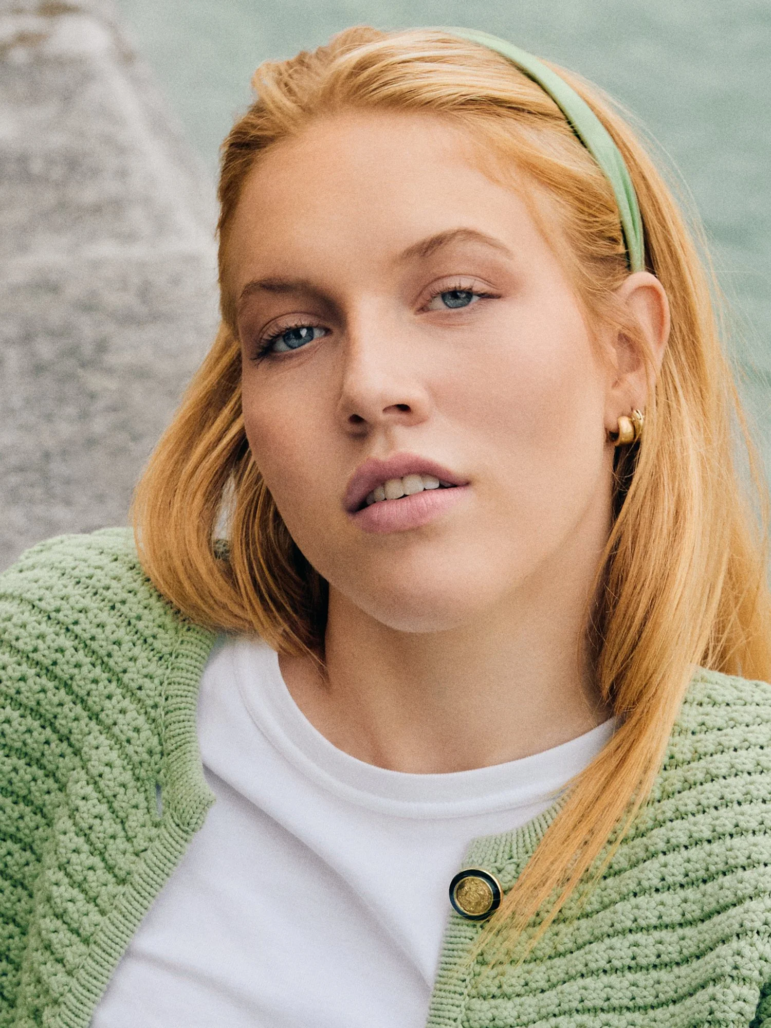 Nahaufnahme eines jungen Mädchens mit roten Haaren, blauen Augen, grüner Haarband und grüner Strickjacke vor Himmel und Wasser.
