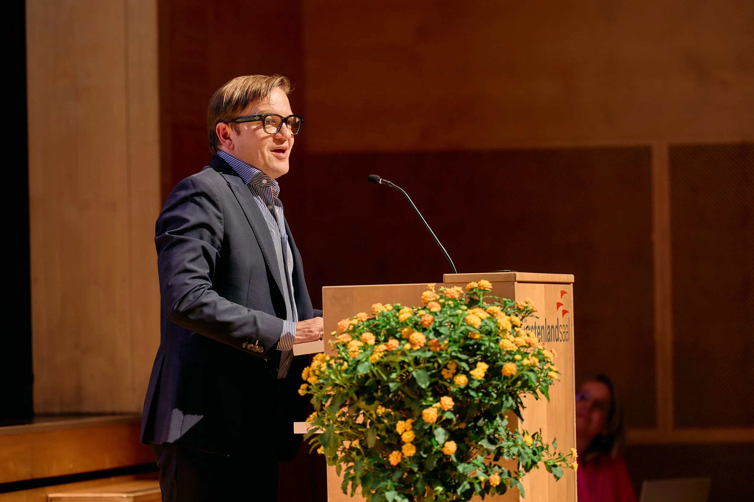 Männlicher Redner auf einer Bühne bei einer Veranstaltung, steht hinter einem Pult mit einem Mikrofon, im Vordergrund Blumen. Hintergrund aus Holzpaneelen.