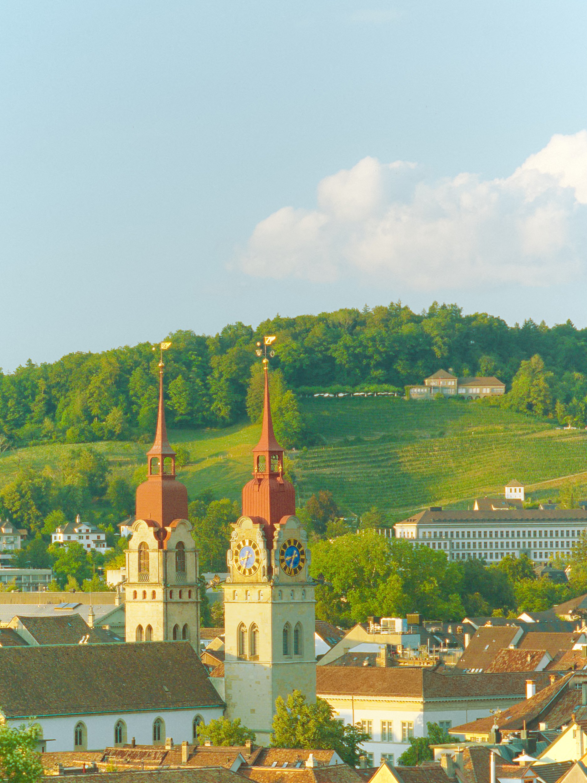 Ansicht einer Stadt mit Kirche, grünen Hügeln im Hintergrund und Himmel mit Wolken.