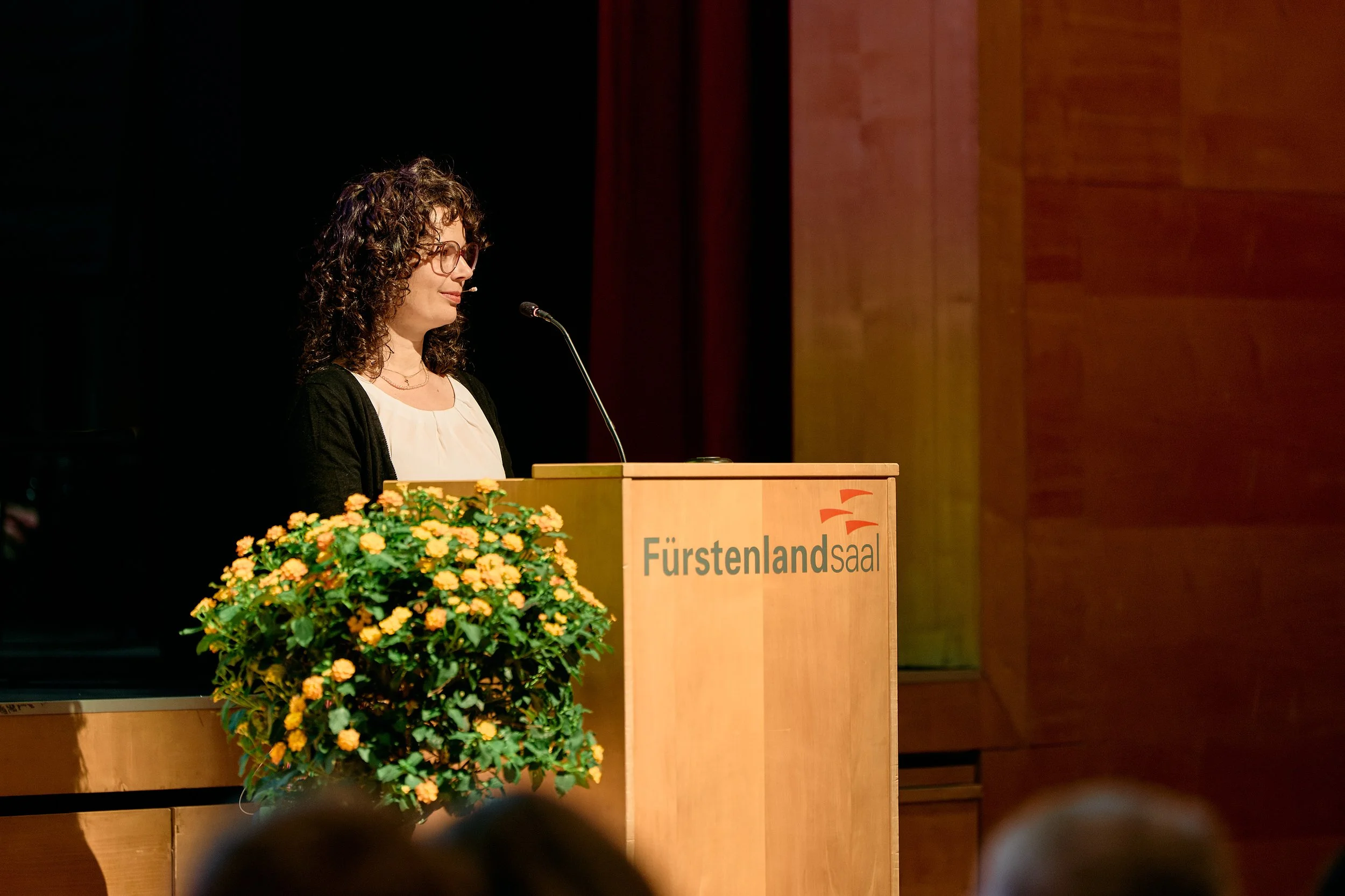 Junge Frau spricht an einem Rednerpult in einem Saal mit Blumen im Vordergrund.