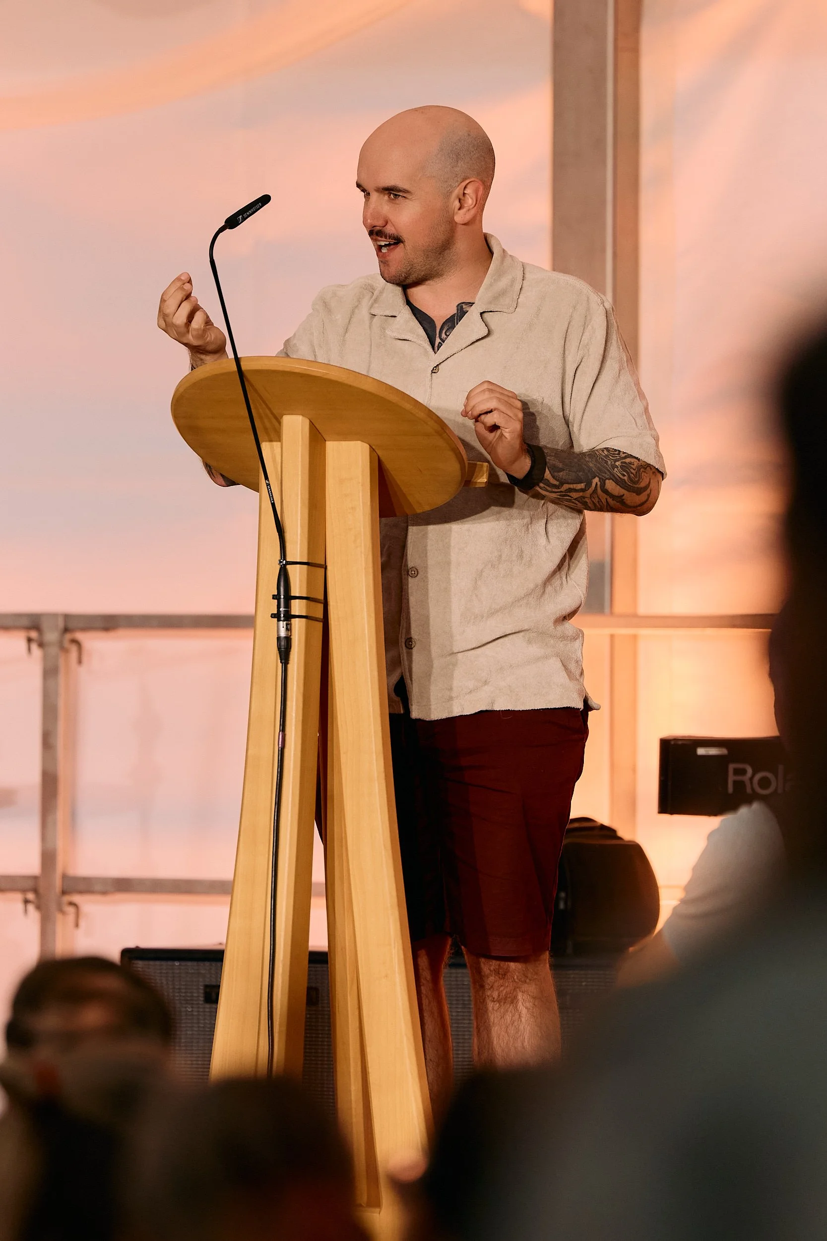 Fotograf & Videograf Wil SG | Businessfotografie Ostschweiz Daniel Dorrer Mann steht an einem Rednerpult aus Holz, spricht in ein Mikrofon, im Hintergrund orangefarbener Himmel. 100 Jahre Gymnasiums Friedberg Gossau SG. Renato Keiser