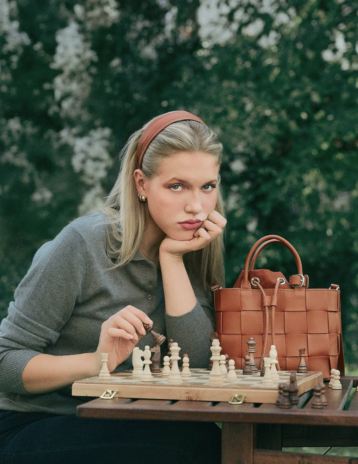 Eine junge Frau mit blonden Haaren, rotbraunem Haarband und grauem Pullover sitzt an einem Schachtisch im Freien und spielt Schach. Sie hat eine nachdenkliche Miene, eine Hand stützt ihr Gesicht, die andere hält eine Schachfigur. Auf dem Tisch liegt 