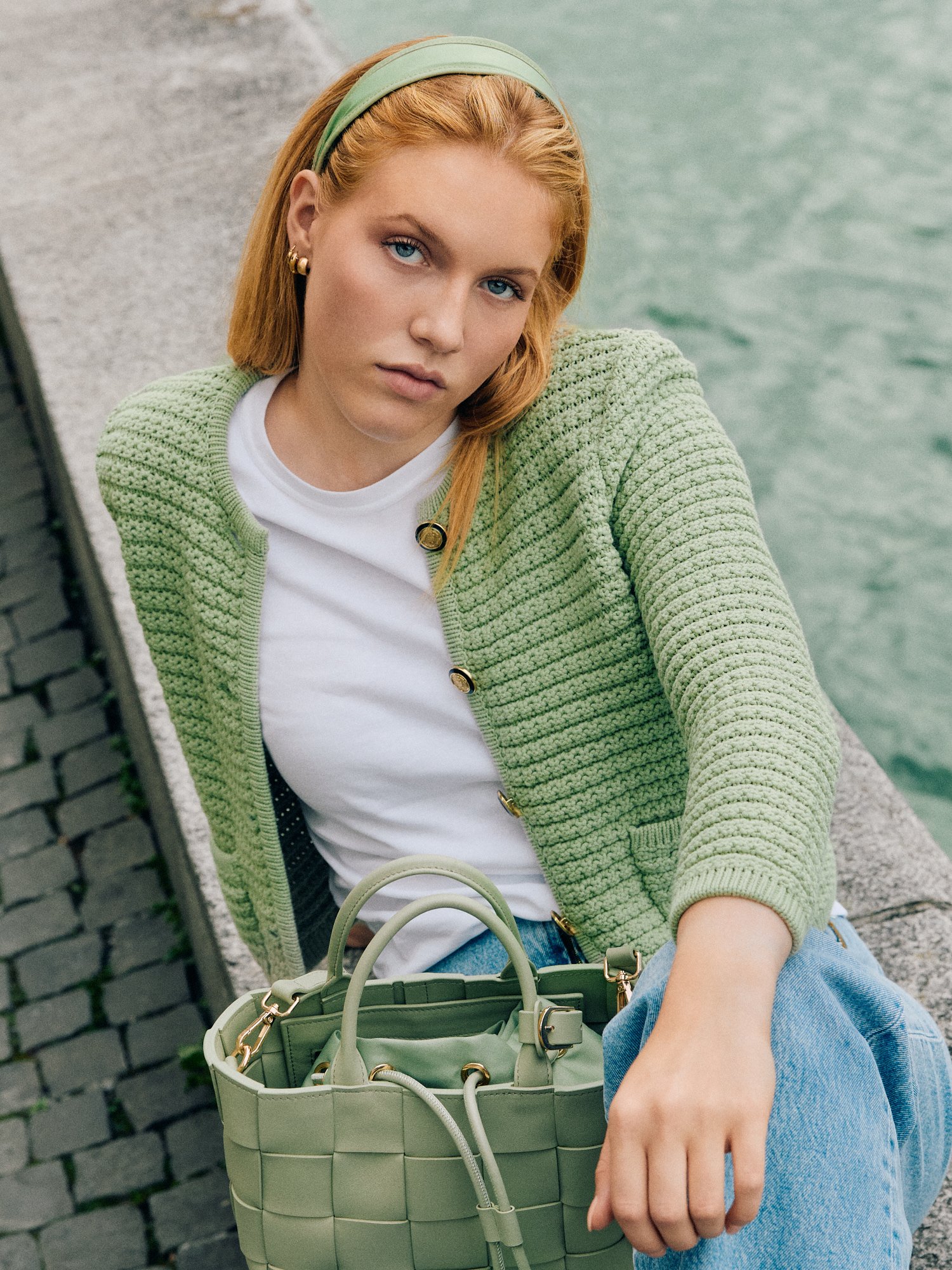 Eine junge Frau mit roten Haaren, blauen Augen und leichten Make-up sitzt auf einer Bank an einem Wasseruf. Sie trägt einen hellgrünen gestrickten Cardigan, ein weißes T-Shirt, eine grüne Handtasche und eine hellgrüne Haarband.