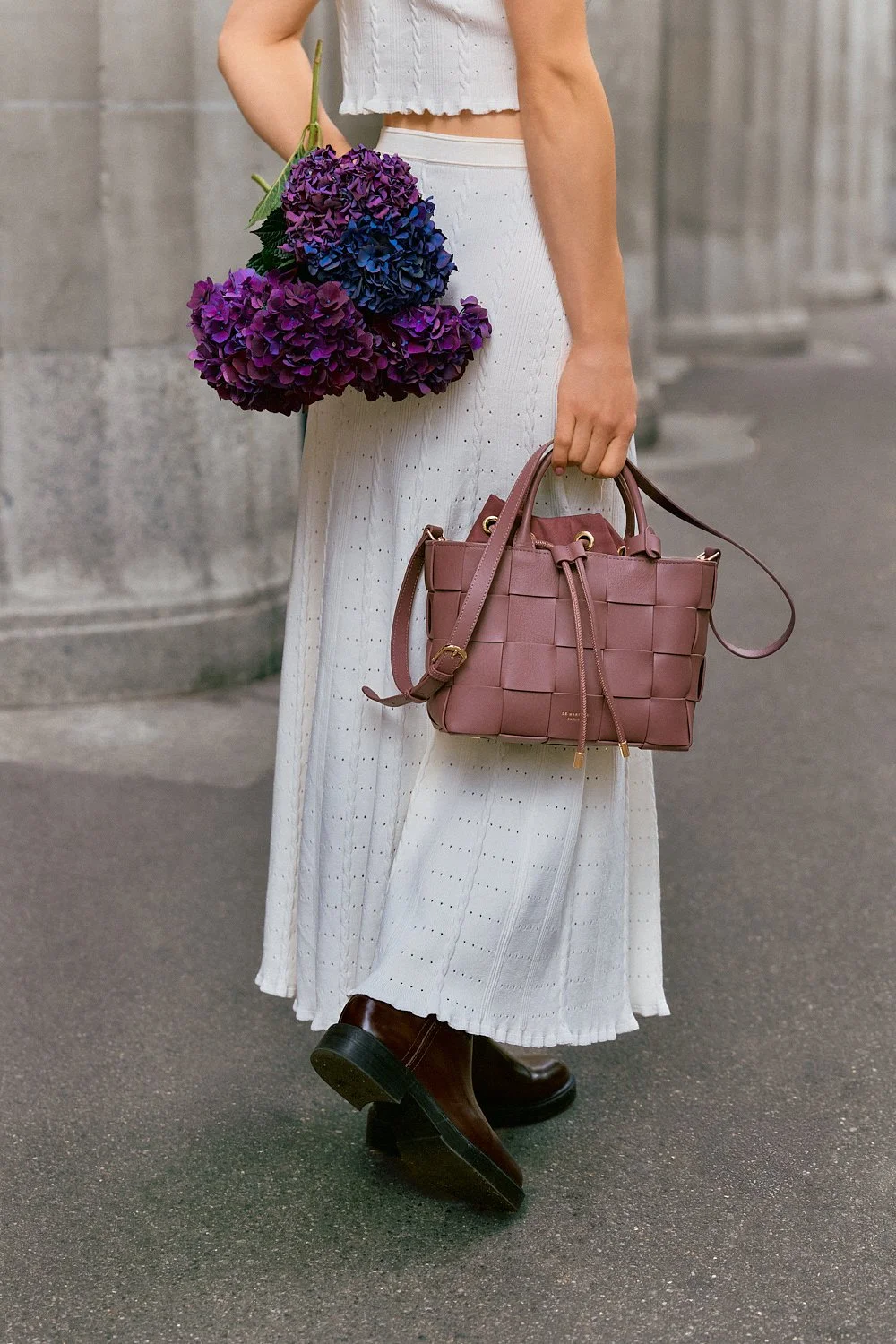 Frau trägt eine weiße, luftige Hose und schwarze Stiefel, hält eine rosa Handtasche und hat einen Bund mit violett-blauen Hortensien. Die Szene findet auf einer Stadtstraße statt.