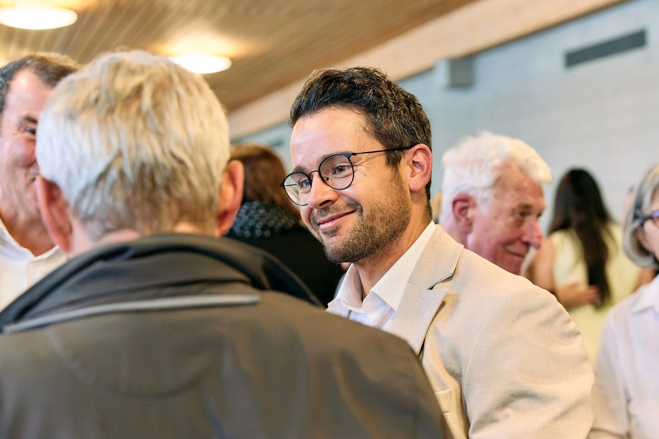 Vier Menschen im Gespräch bei einer Veranstaltung, einer trägt eine Brille und weißes Hemd, weitere Personen im Hintergrund.