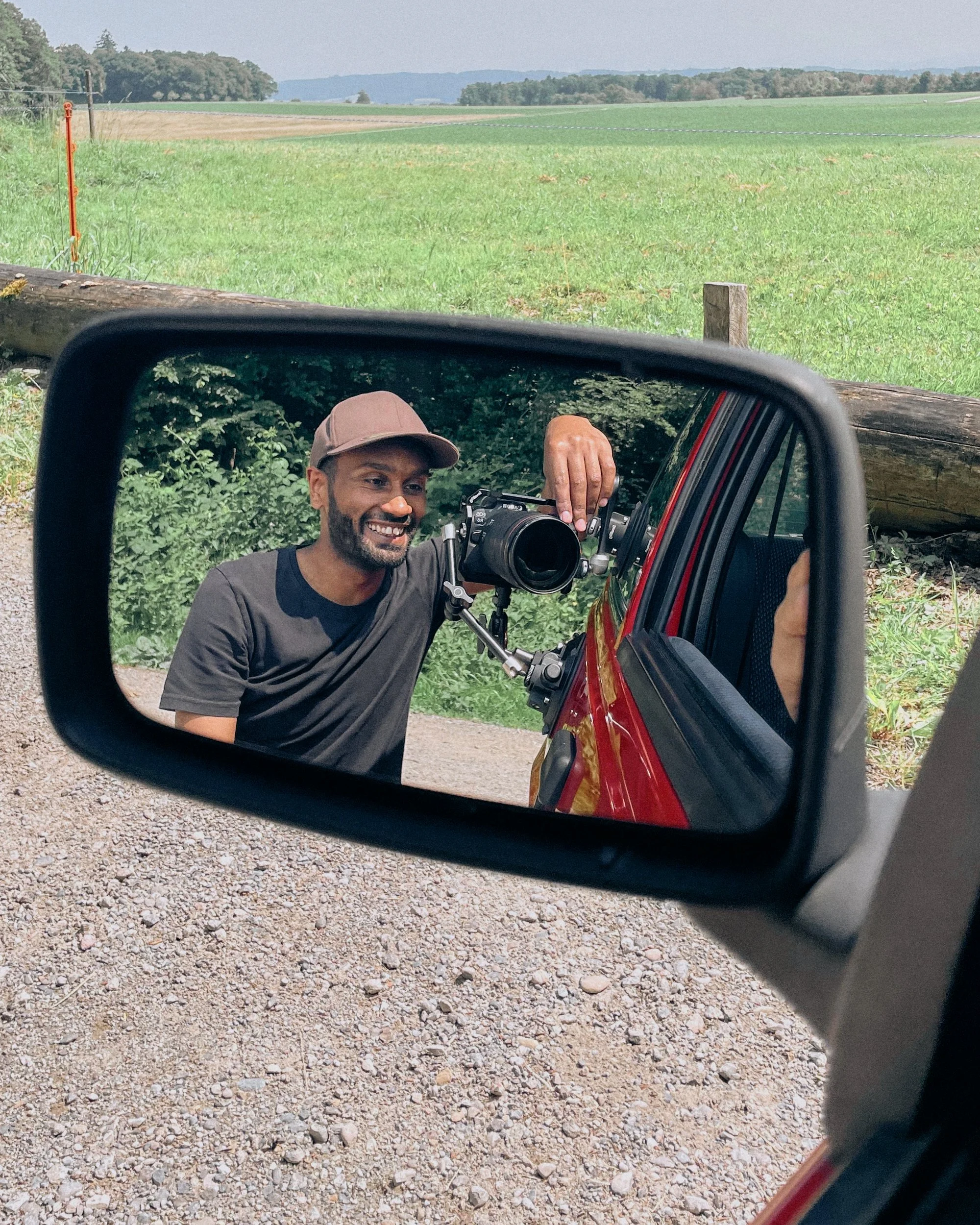 Ein Mann macht ein Selfie im Seitenspiegel eines roten Fahrzeugs, im Hintergrund eine ländliche Landschaft mit grünen Feldern und Bäumen.
