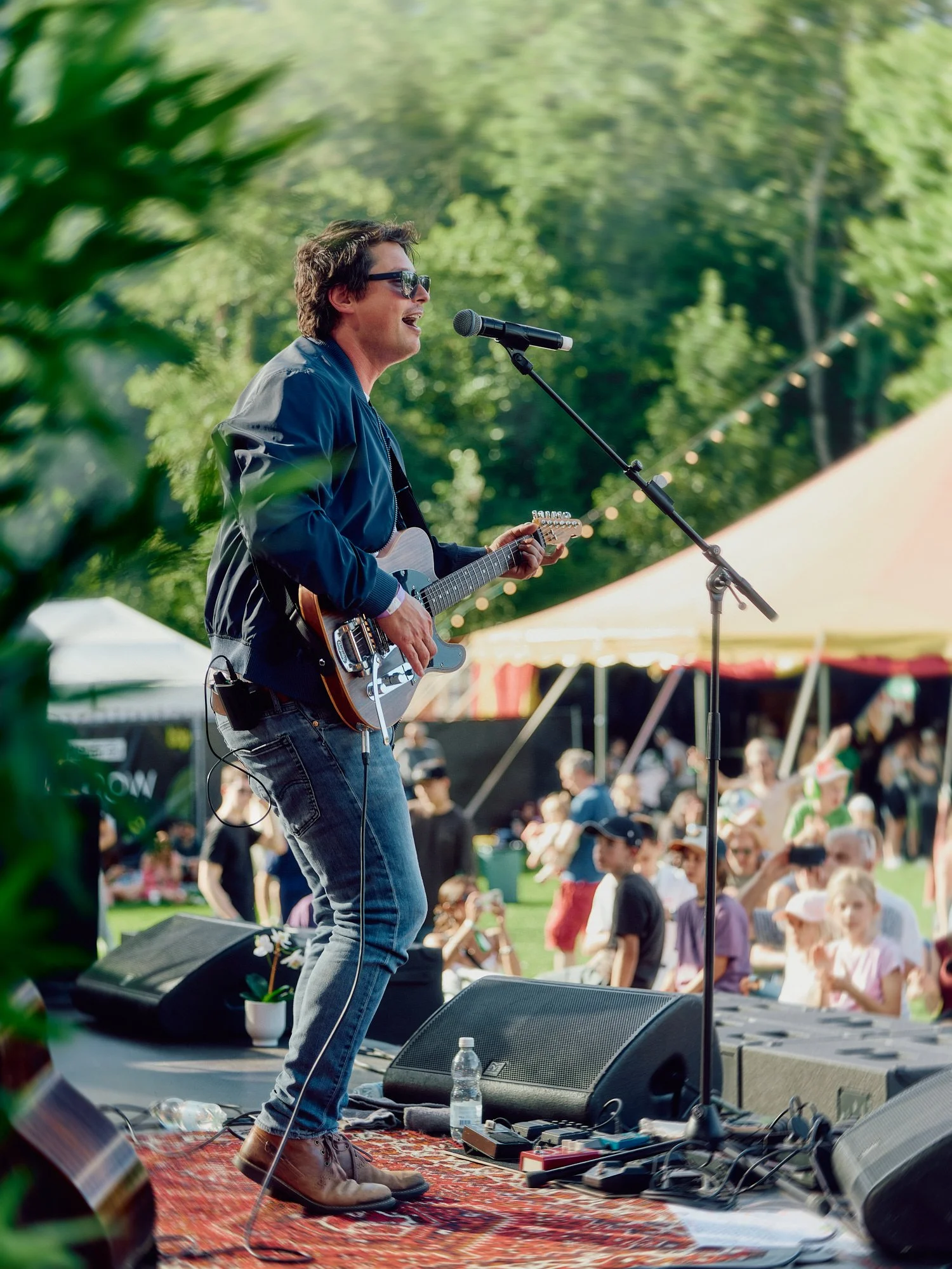 Ein junger Mann spielt Gitarre auf einer Bühne in einem Park mit grünem Laub im Hintergrund, während ein Publikum im Freien zusieht. Daniel Dorrer Film und Bild