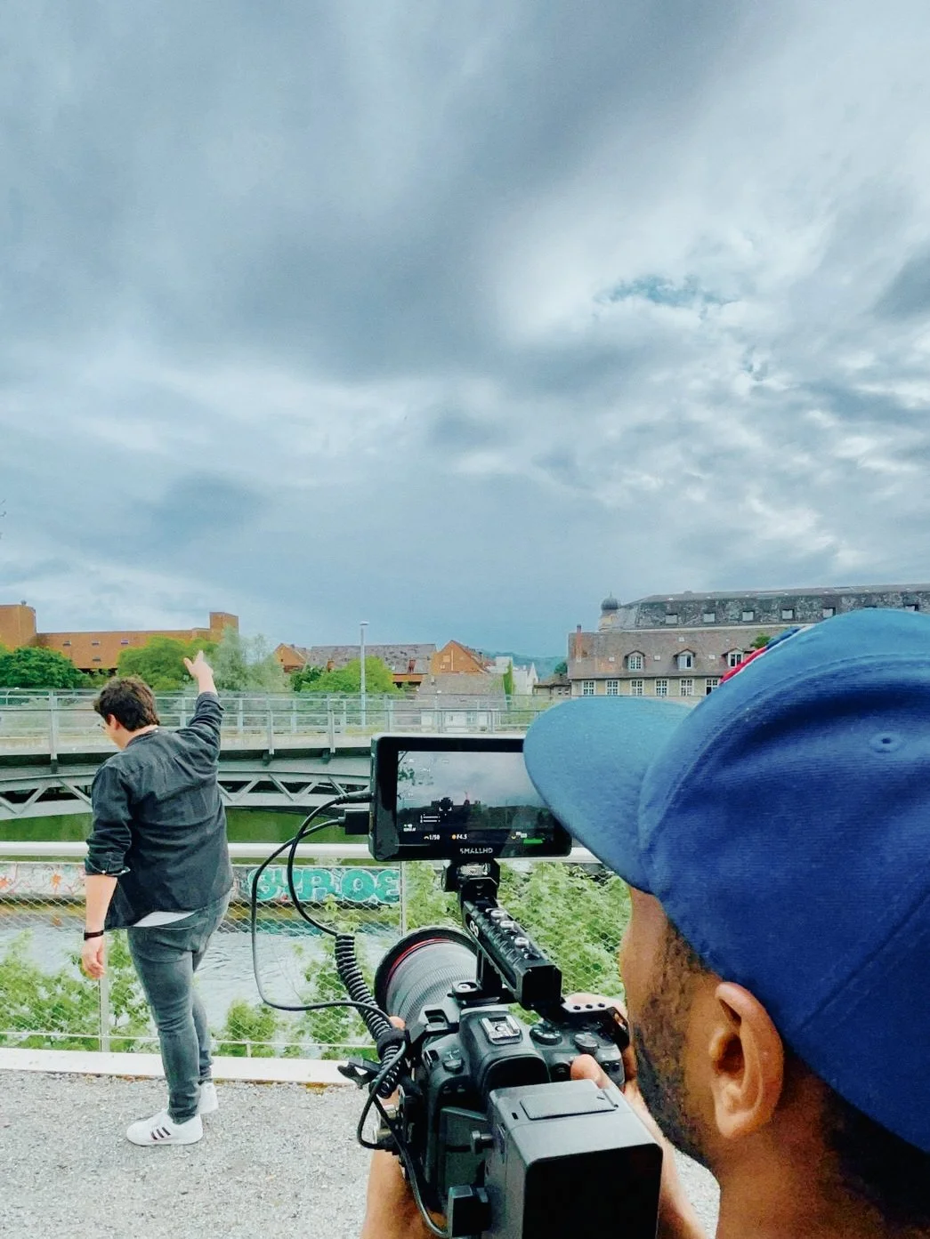Ein Mann mit blauer Cap filmt eine Person, die auf einer Brücke steht und nach oben zeigt, bei bewölktem Himmel.
