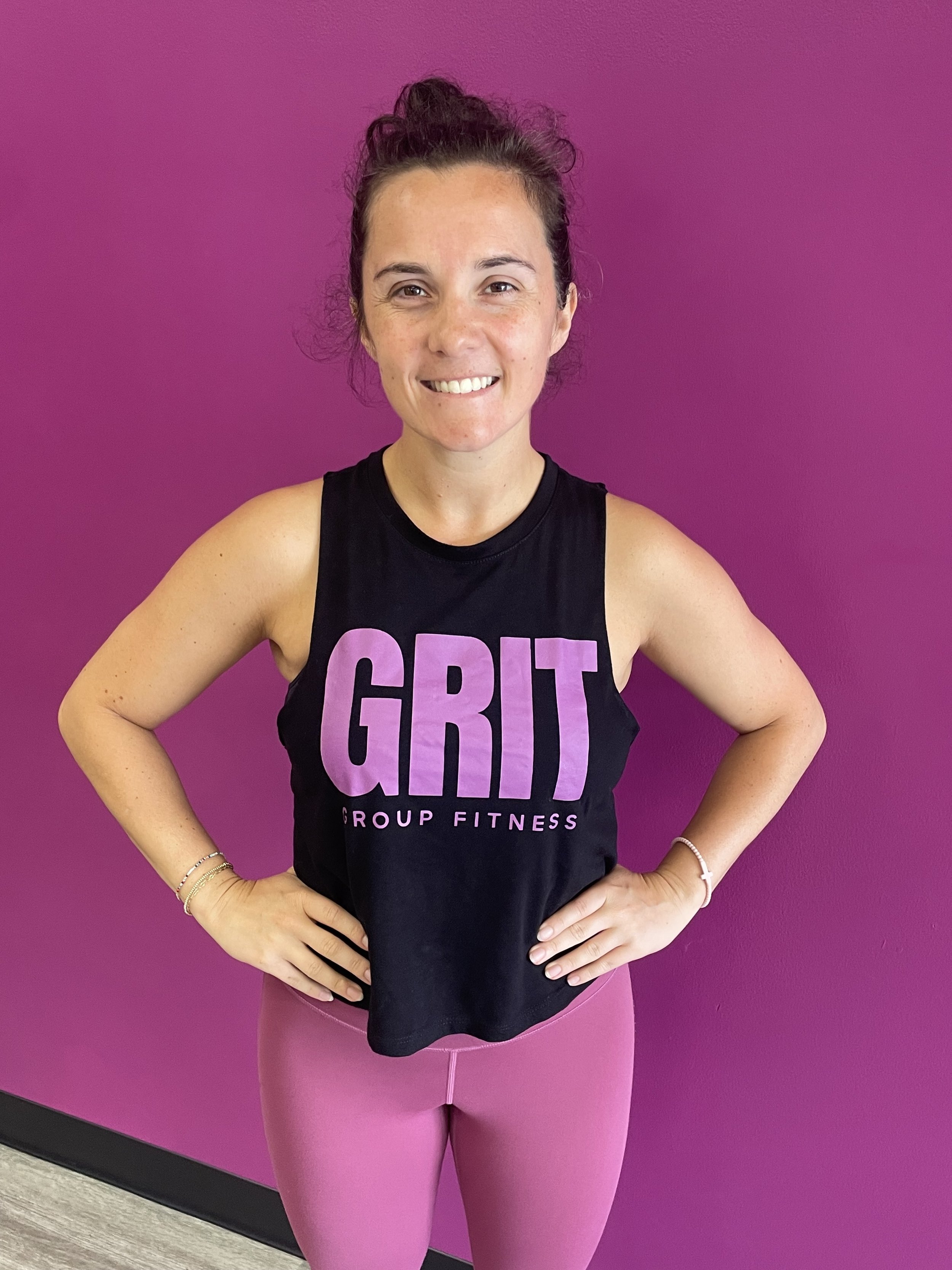 A woman with curly hair in a high bun, smiling, wearing a black tank top with the word 'GRIT' in large pink letters, and pink leggings, standing against a purple wall in a fitness studio.