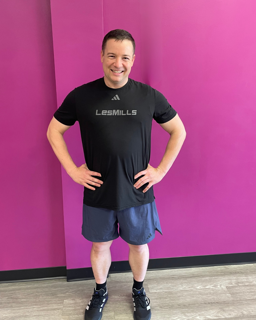 A man standing with hands on hips in front of a pink wall, wearing a black Adidas Les Mills t-shirt, dark shorts, and athletic shoes, smiling at the camera.