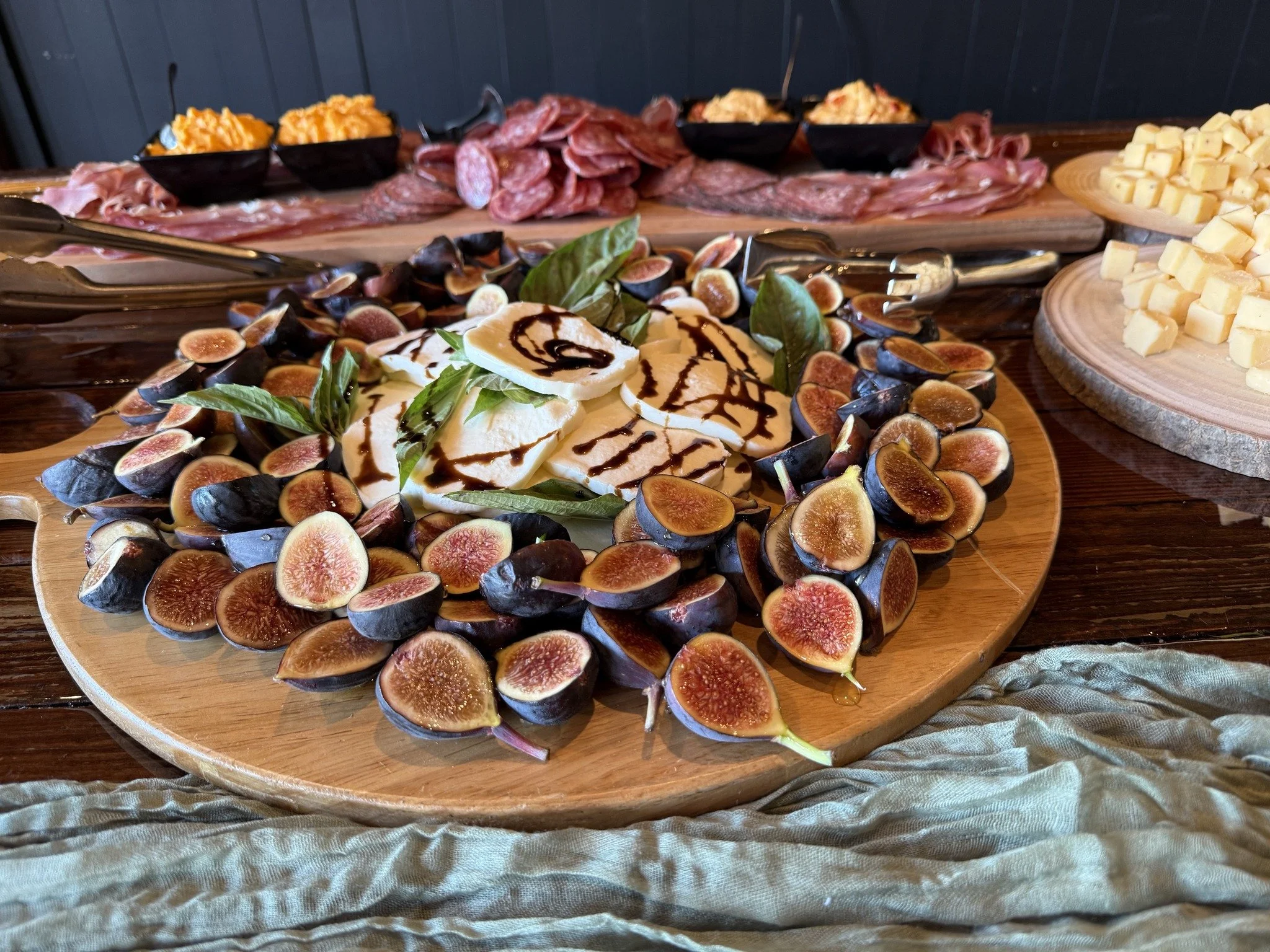 A platter of sliced mozzarella cheese with balsamic glaze and fresh basil, surrounded by fresh figs on a wooden serving board, with a charcuterie spread of cured meats, cheeses, and accompaniments in the background.
