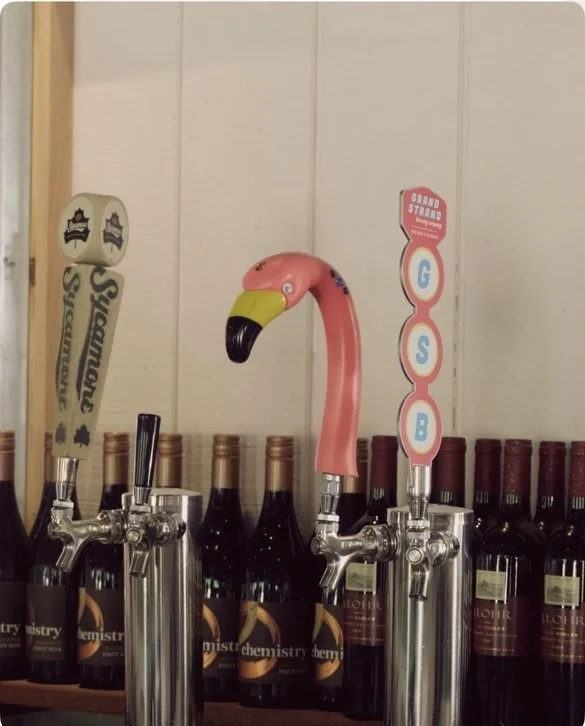 Beer tap handles, including one shaped like a flamingo, on a bar counter with bottles of wine in the background.