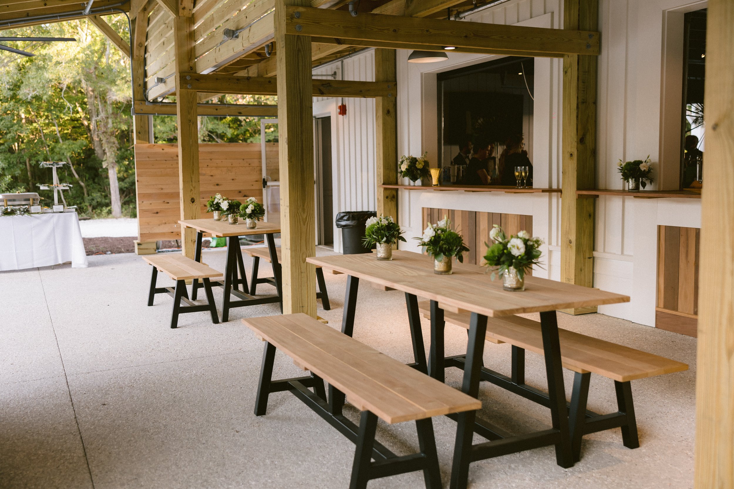 Outdoor patio with wooden picnic tables, decorated with green and white floral arrangements, beneath a wooden canopy.