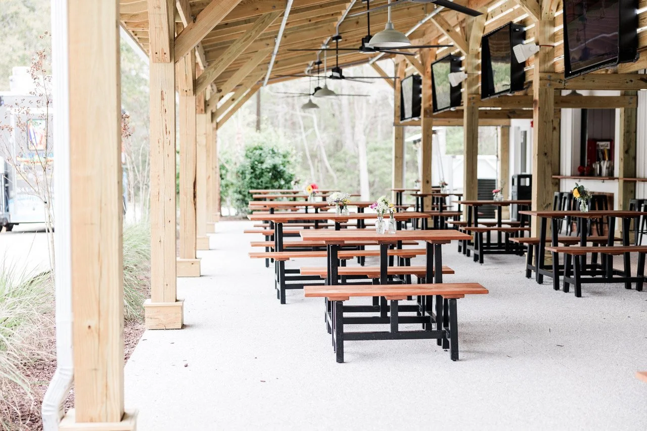Empty outdoor dining area with wooden tables and benches, decorated with small flower vases, and a wooden pergola structure over the space with hanging lights and TV screens on the wall.