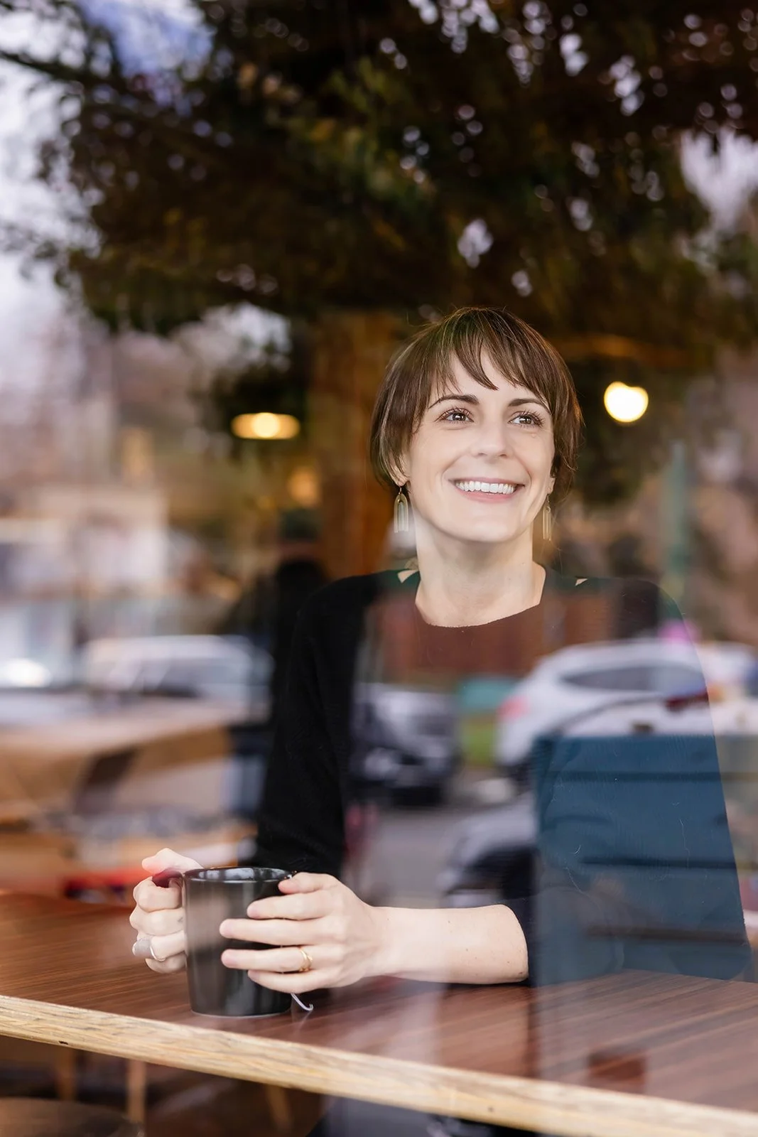 Woman with short brown hair and earrings smiling while sitting at a table in a cafe, holding a black mug, seen through a window with reflections of parked cars and a tree outside.