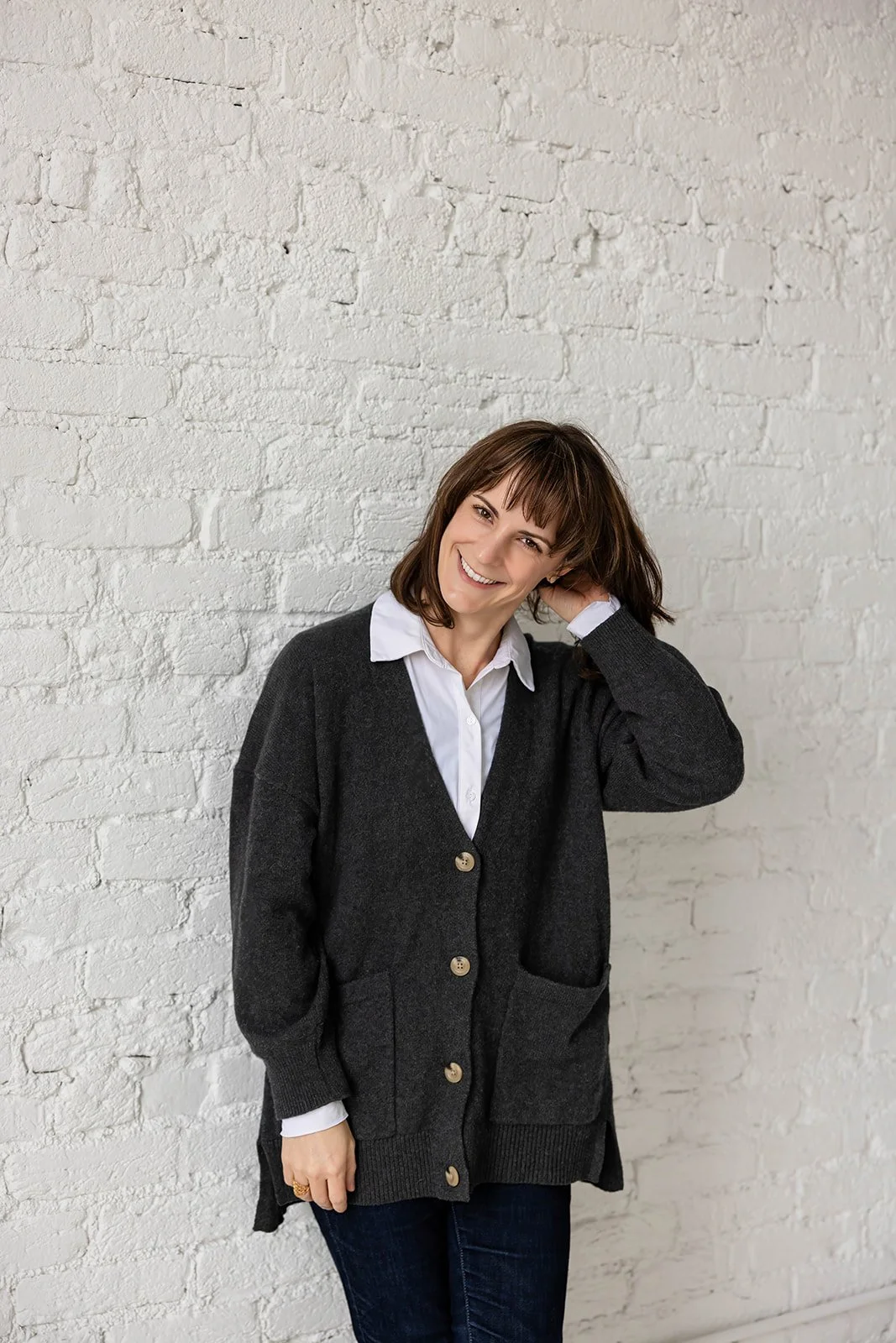 A woman with brown hair, wearing a white collared shirt and a dark gray cardigan, smiling and touching her hair, standing against a white brick wall.