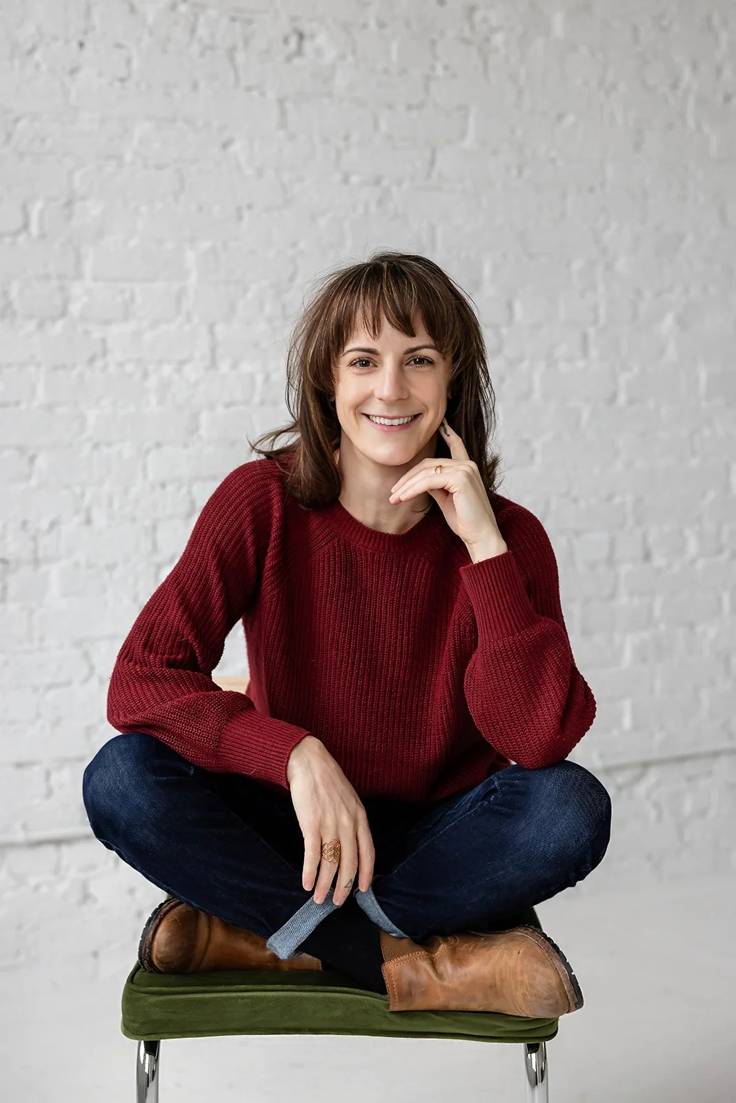 A woman with shoulder-length brown hair, wearing a red sweater, dark jeans, and brown boots, is sitting cross-legged on a green chair in front of a white brick wall, smiling at the camera.