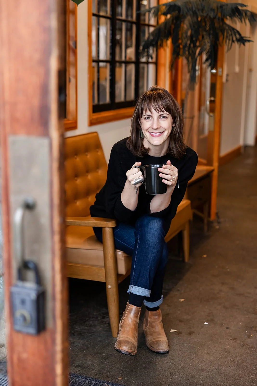 Woman sitting on a wooden bench inside a coffee shop, holding a black mug and smiling at the camera.