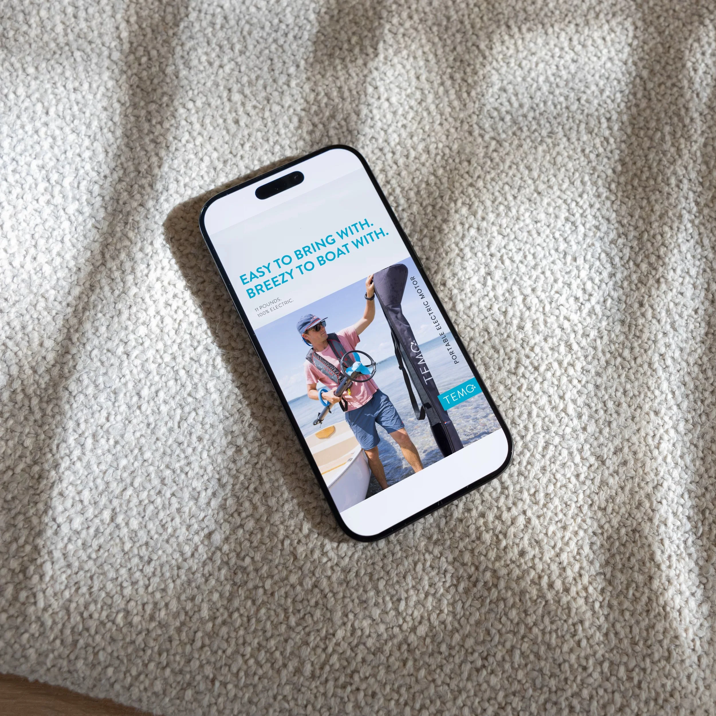 Smartphone displaying an advertisement for portable electric motors for boating, featuring a man on a boat holding a shadowed electric motor, on a textured beige surface.