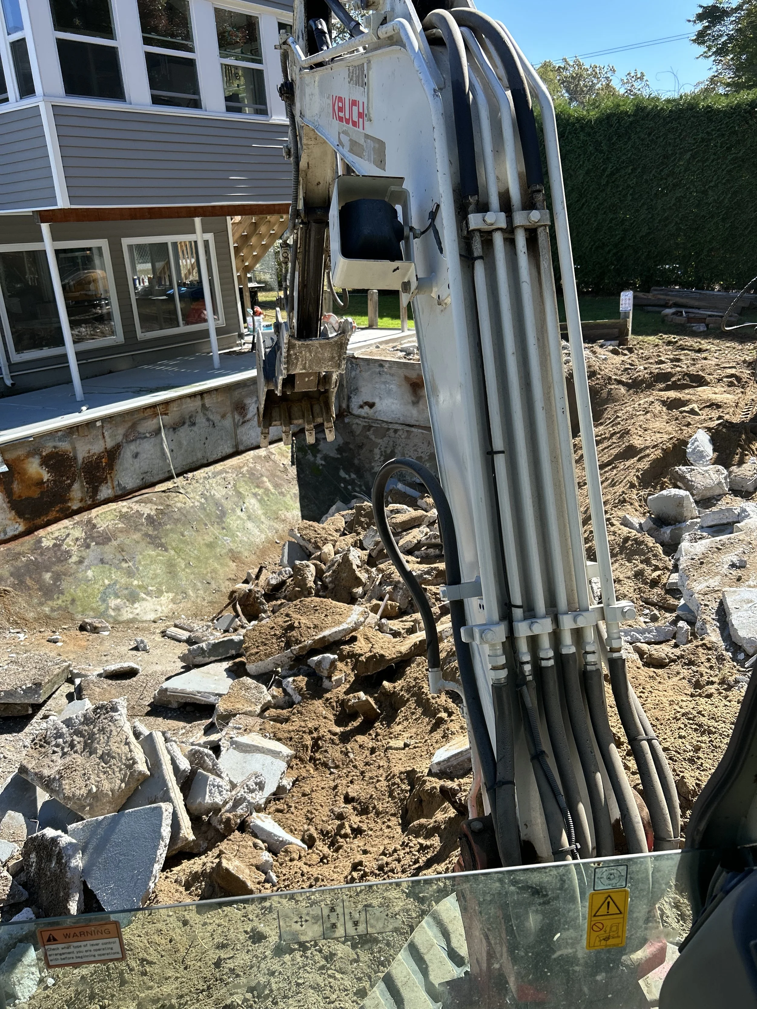 View from inside a construction excavator showing the arm and bucket digging into dirt and rubble at a construction site, with a house and trees in the background.