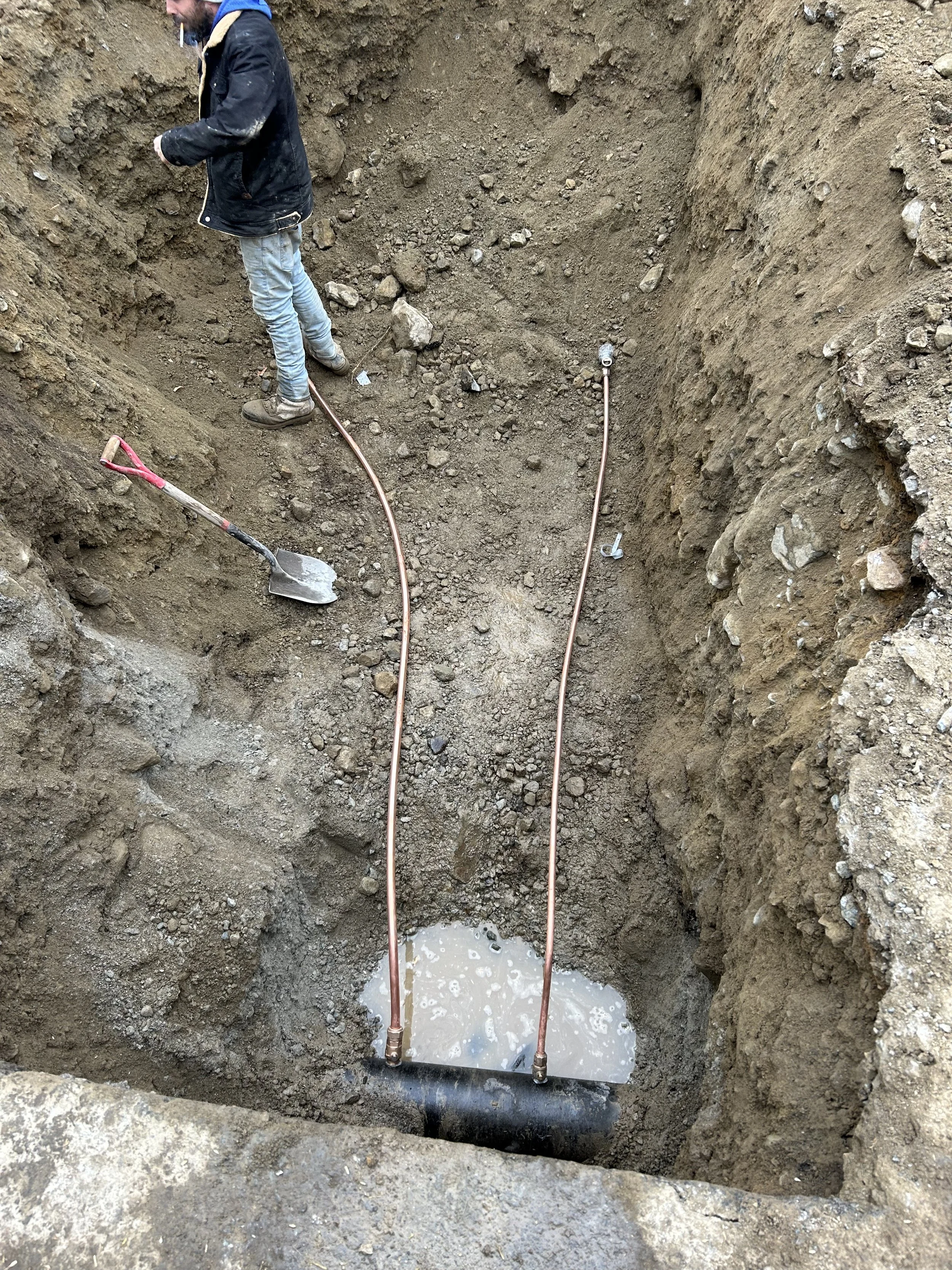 A person working in a trench with copper pipes, water, and construction tools.
