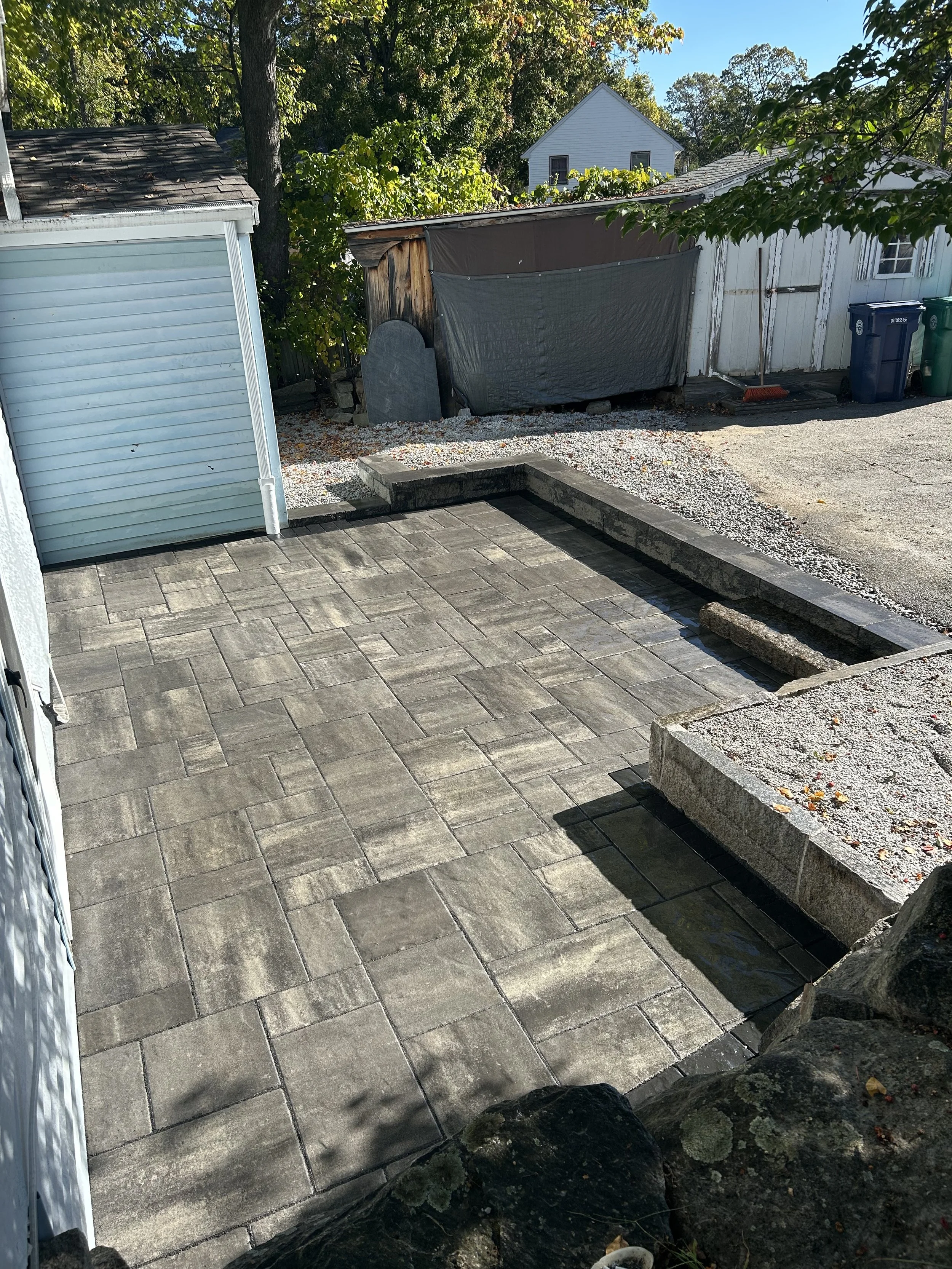 Backyard patio with new stone paving, a small raised bed with gravel, and a shed with other storage structures and trash bins in the background.