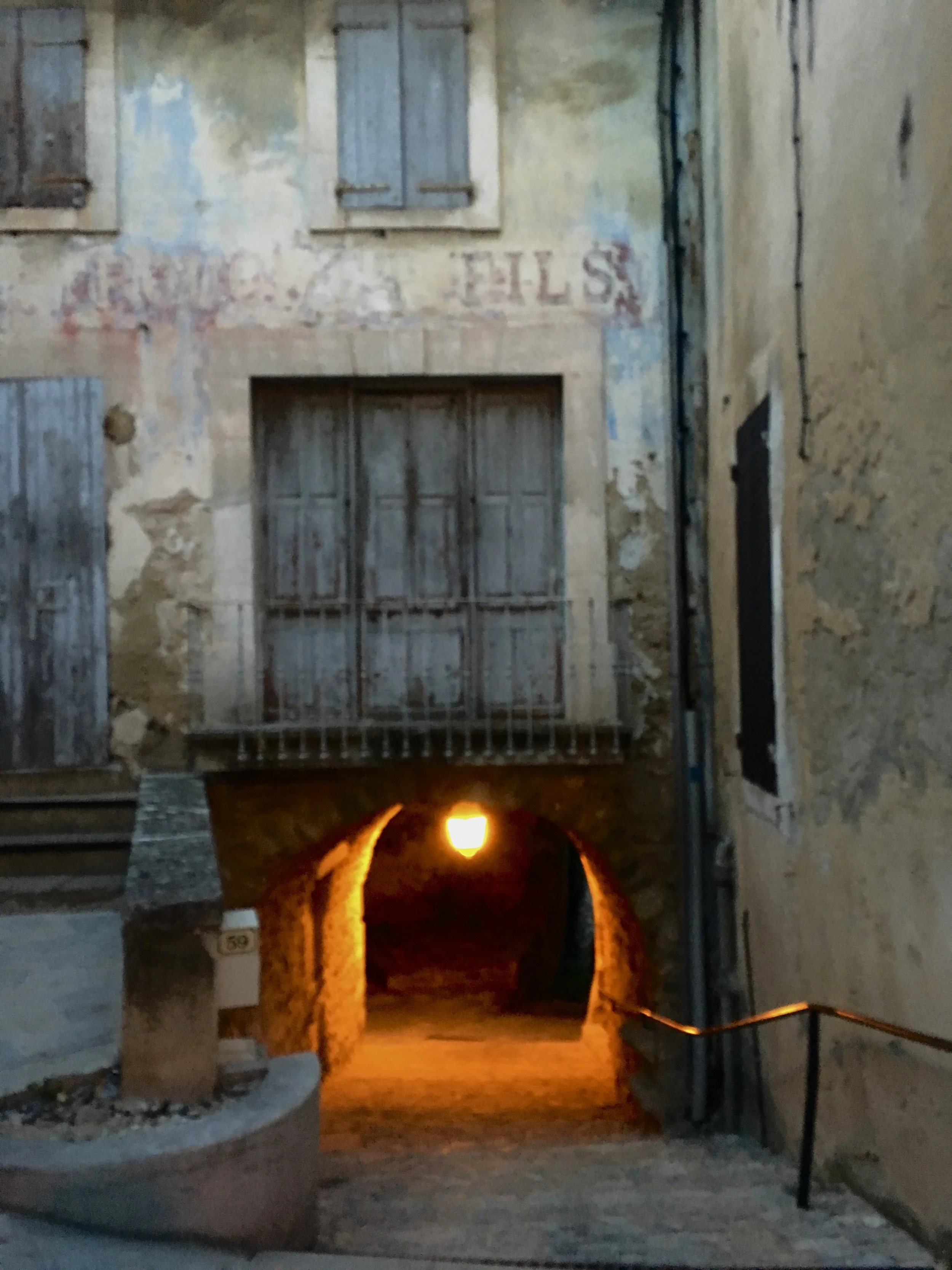 A small tunnel with stone walls and an orange glow from a light inside, leading into a dark alley, with old weathered buildings on either side.