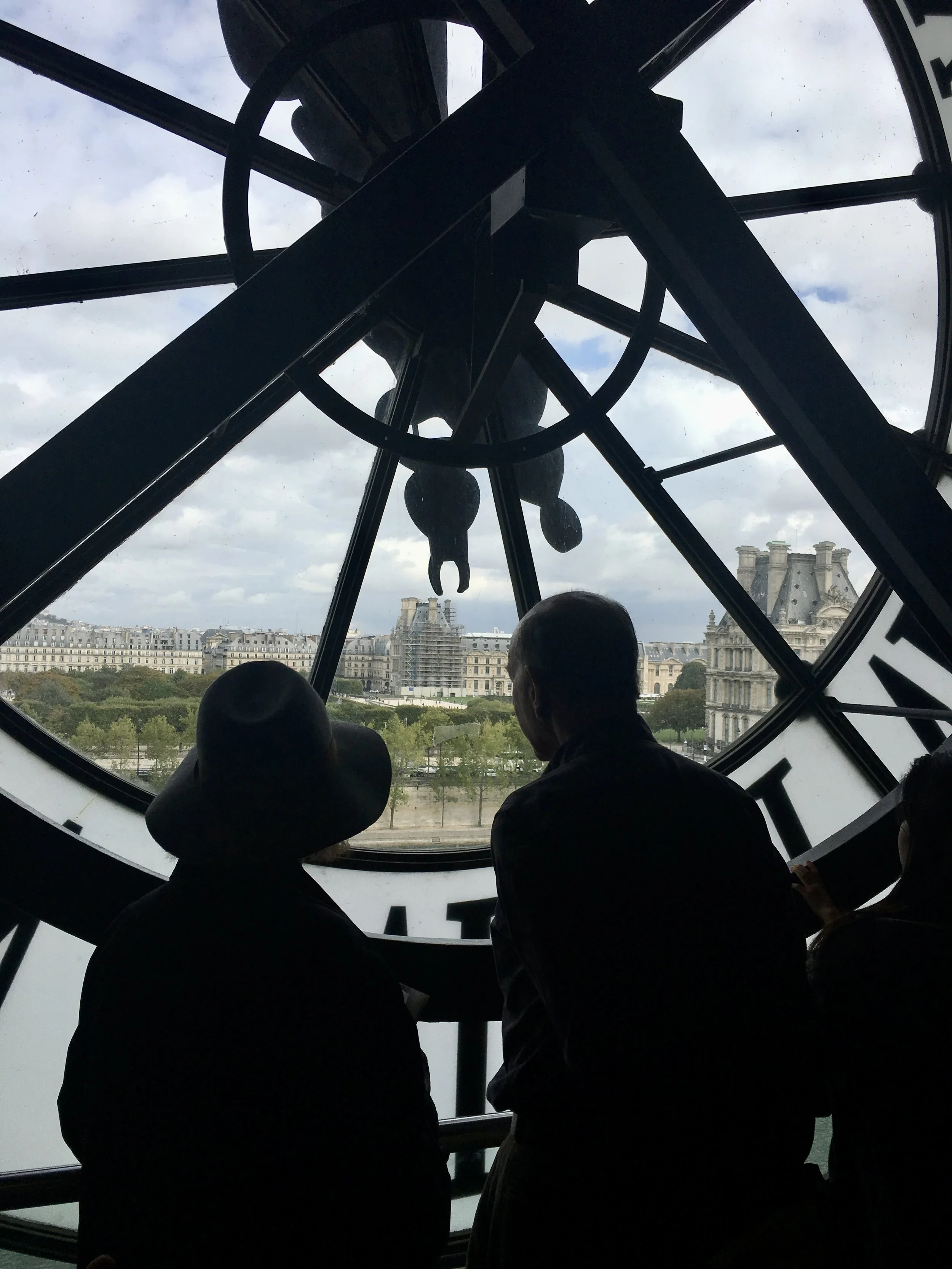 View from inside the Musée d'Orsay clock window with three silhouetted people looking out at Paris, including the Sacré-Cœur Basilica visible in the distance.